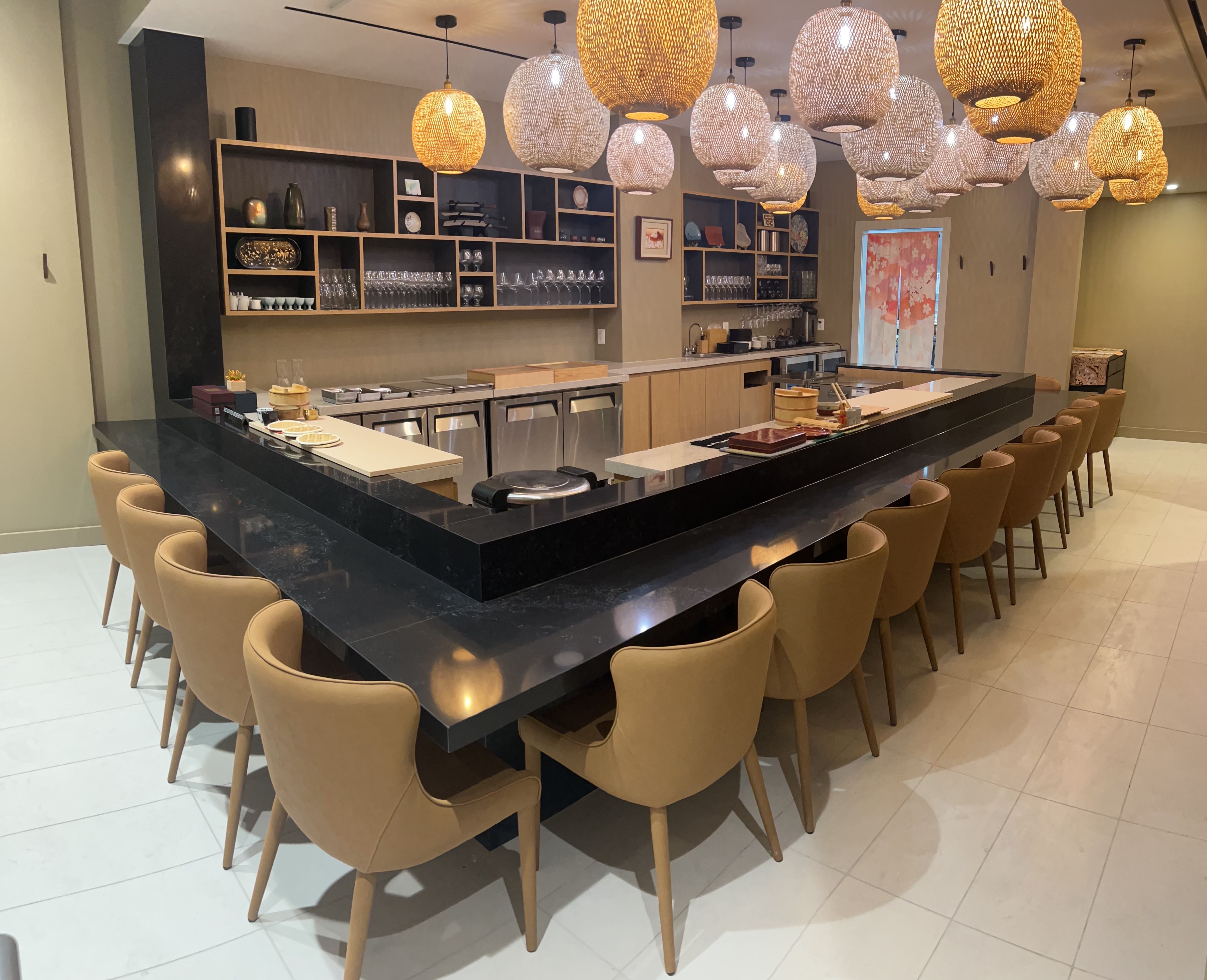 Modern sushi bar with a long black marble counter and tan stools around it. Warm woven pendant lights, glassware shelves behind, and a pink blossom door at the back.