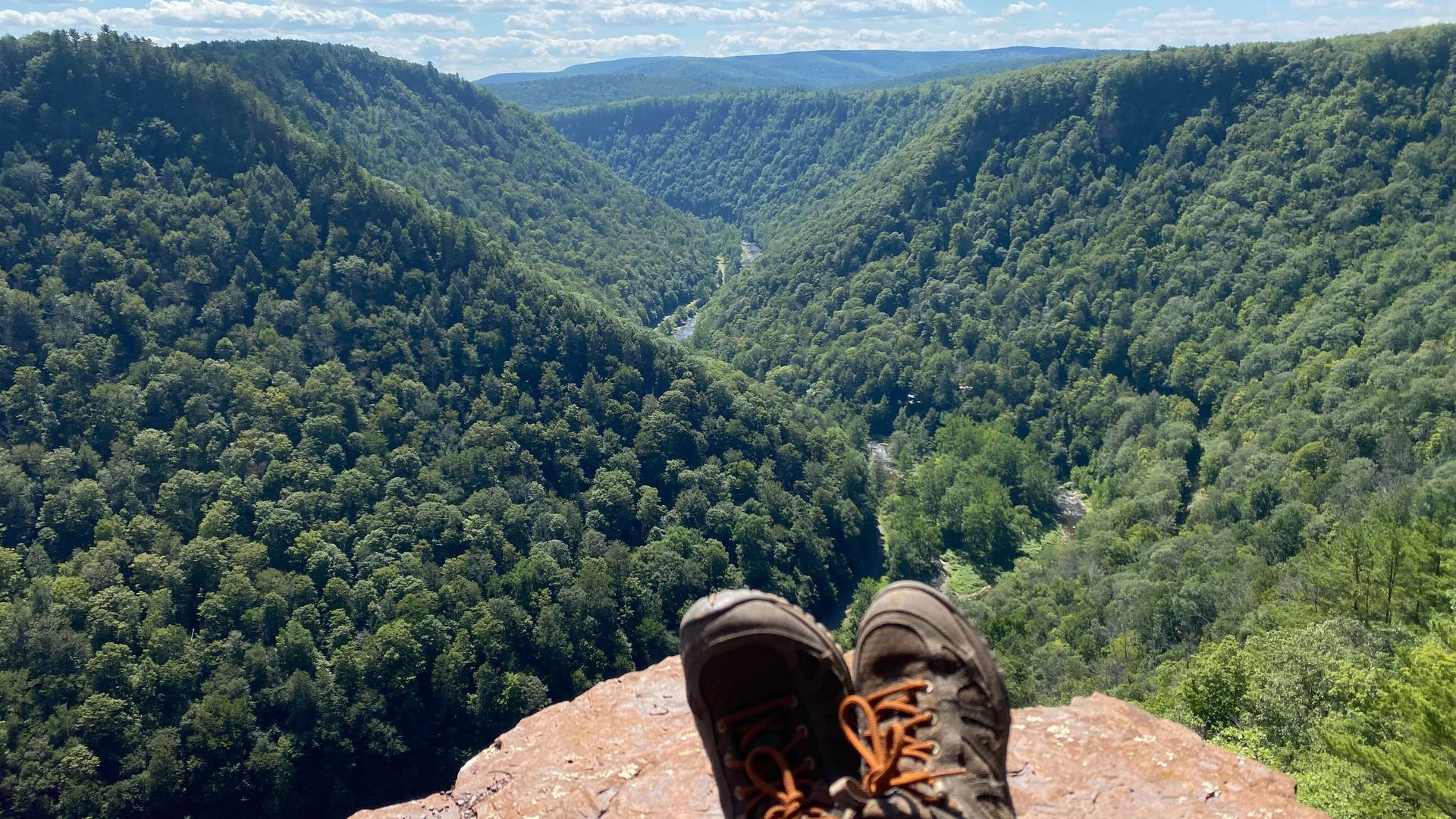View of a deep, green forested valley with a river running through it, seen from a rocky cliff where a person’s legs and brown hiking shoes with orange laces are visible. Clear blue sky with clouds.