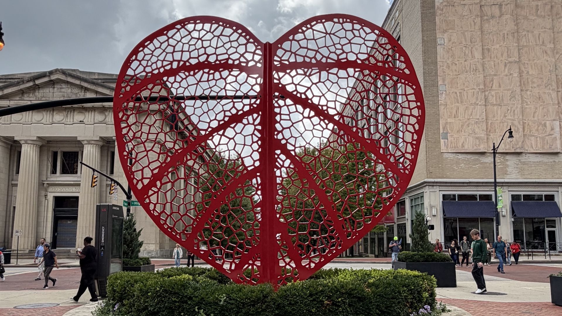 Large red open-work sculpture shaped like a heart, with a honeycomb lattice, set in a circular brick planter in a city square; historic building facades and pedestrians in the background.