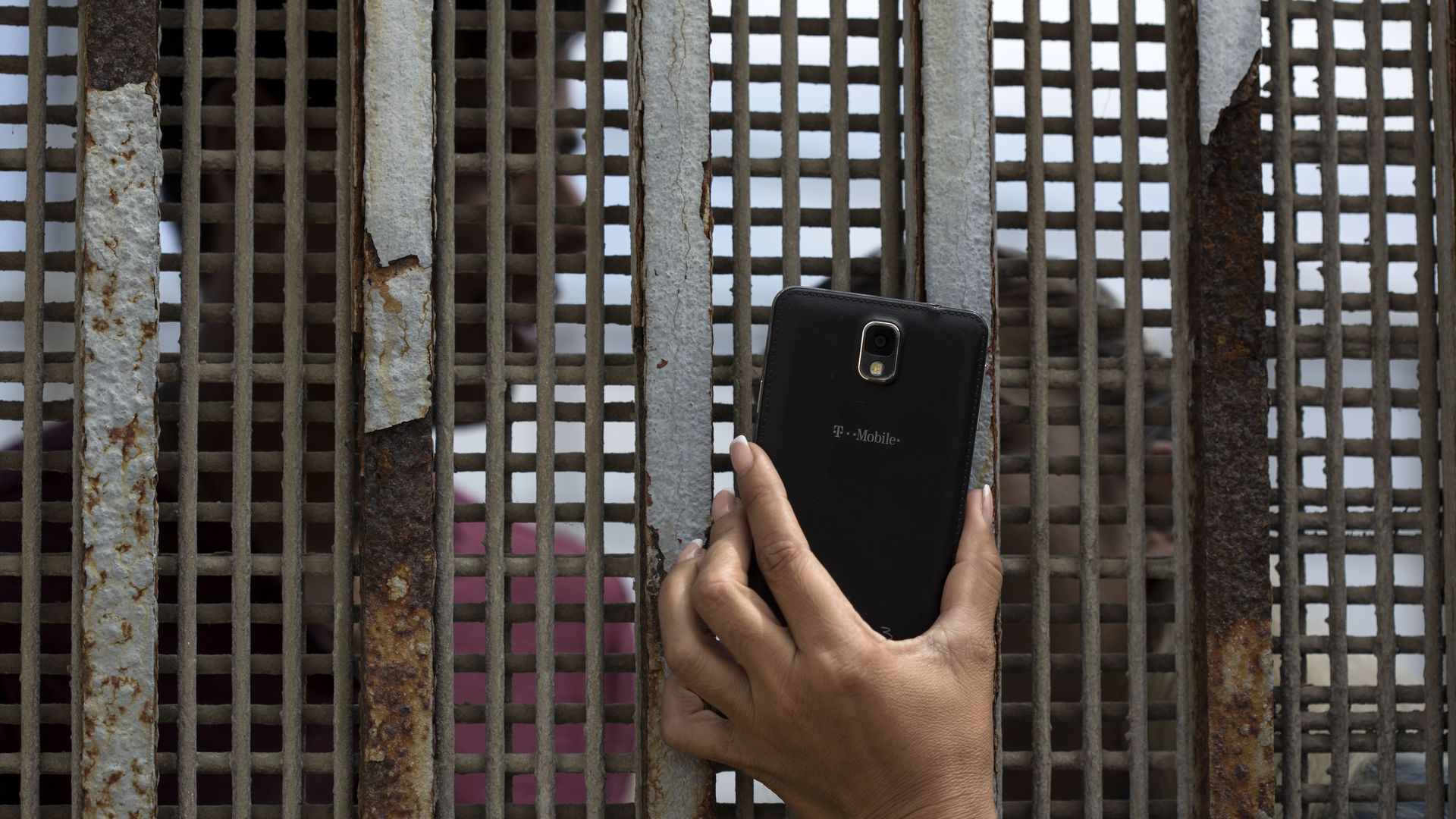 A hand holds a phone up to a grated border wall.