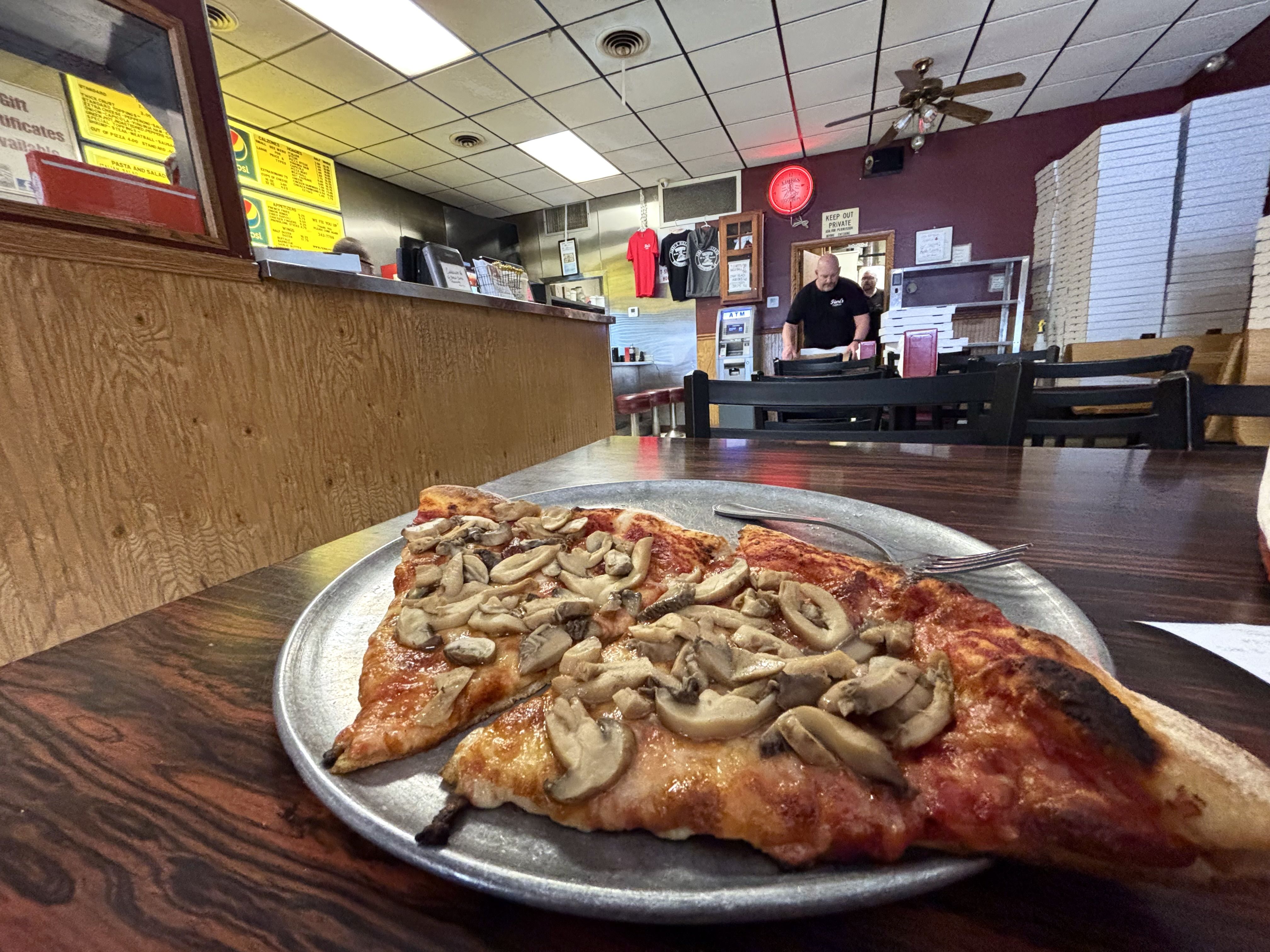 Foreground shows a large mushroom-topped pizza on a metal tray, on a dark wooden table inside a casual pizza shop. In the background, a bald man in a black shirt stands at the counter near yellow menu boards.