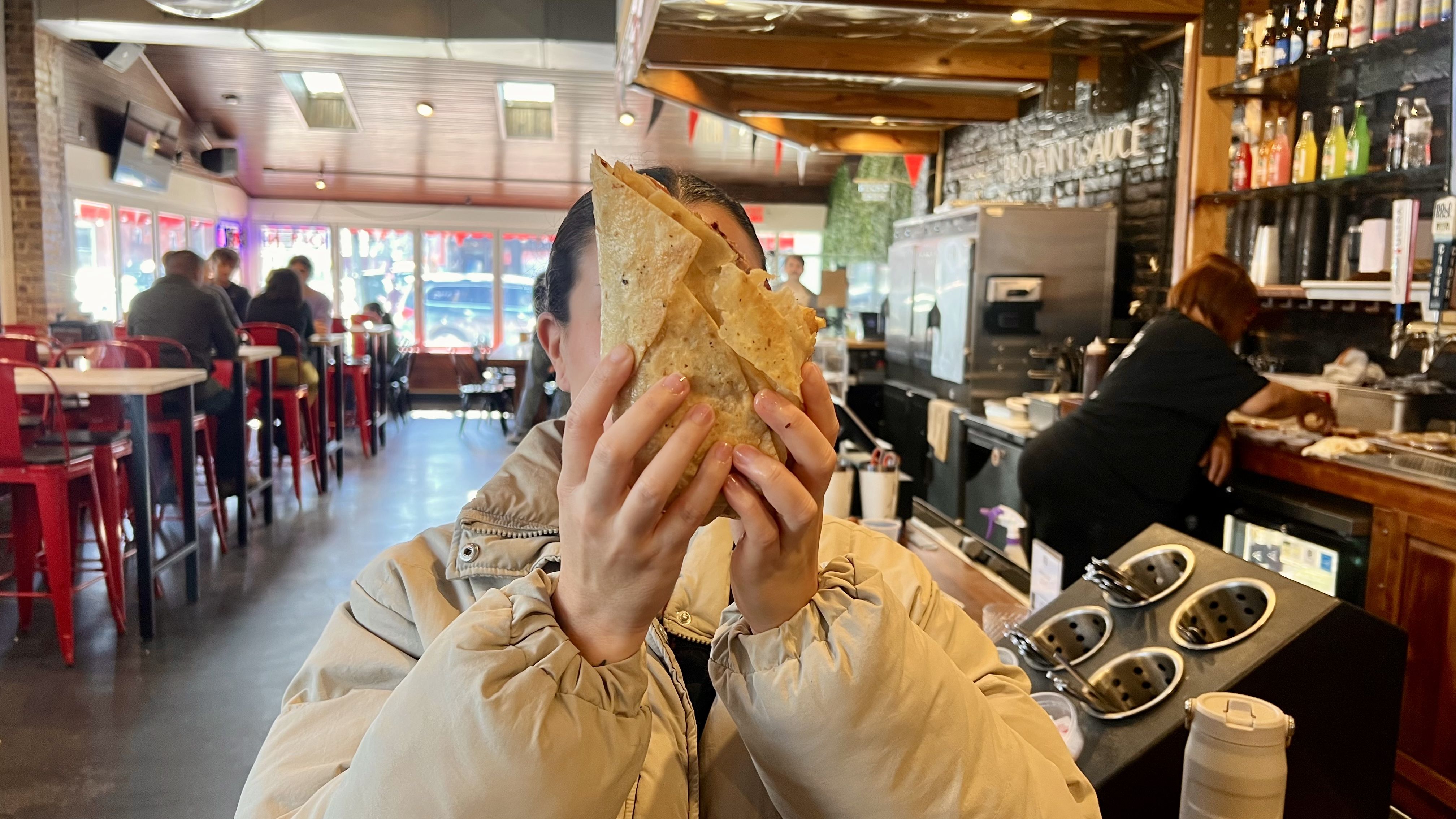 Person in light beige jacket holding a large folded tortilla in front of their face inside a casual restaurant with red stools and a busy kitchen area in the background.