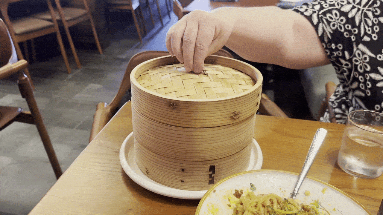 Person in a black-and-white patterned top places a lid on a stacked bamboo steamer basket at a restaurant table; a plate of noodles with a spoon and a glass of water are nearby.