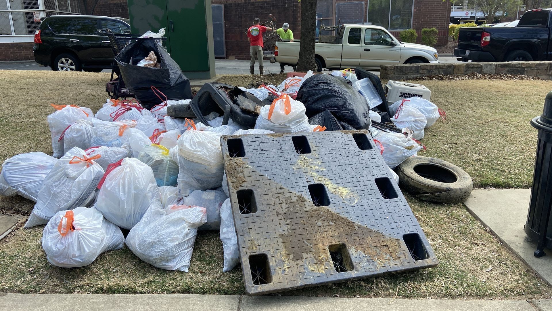 Pile of white trash bags with orange ties and a large black bag on a grassy area beside a sidewalk; a diamond-plate pallet and a tire lie among debris, with two people and vehicles in the background.