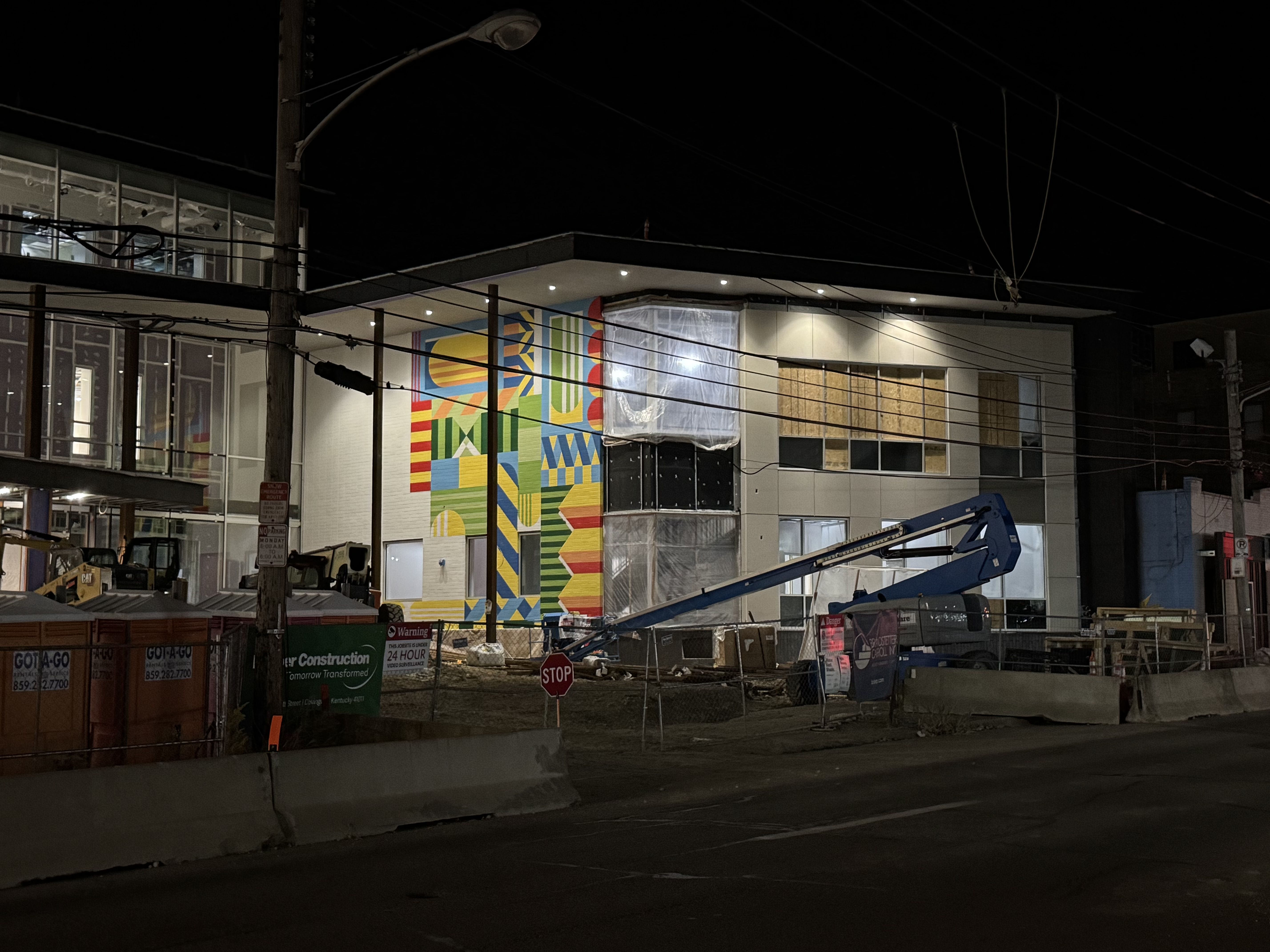 Nighttime construction site with a modern building featuring a large colorful geometric mural, glass windows, and construction equipment. Power lines crisscross the foreground.