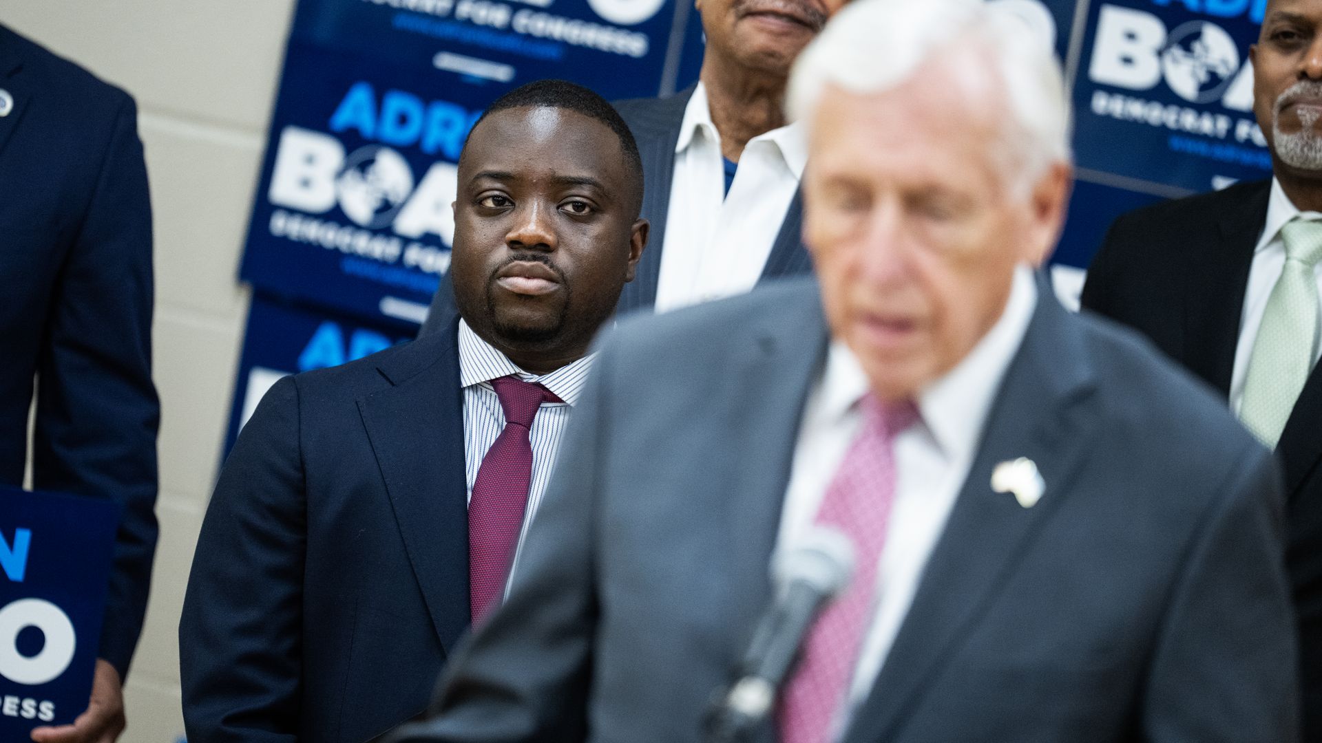 Portrait of a man in a dark suit and maroon tie at a press event, with blue campaign banners in the background. A blurred white-haired man in a gray suit sits near the foreground.