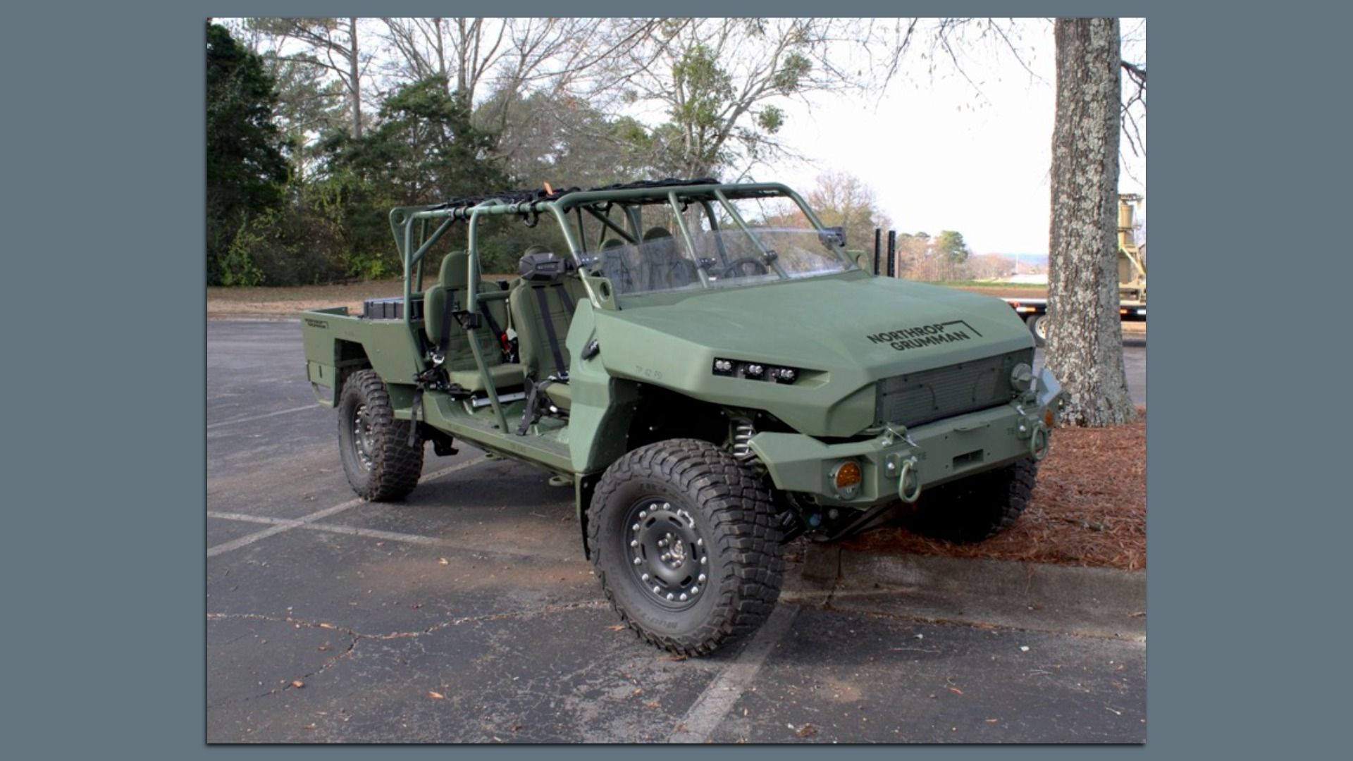 Green open-frame military vehicle with four seats and rugged tires, parked on asphalt near trees. "Northrop Grumman" logo on the front hood.
