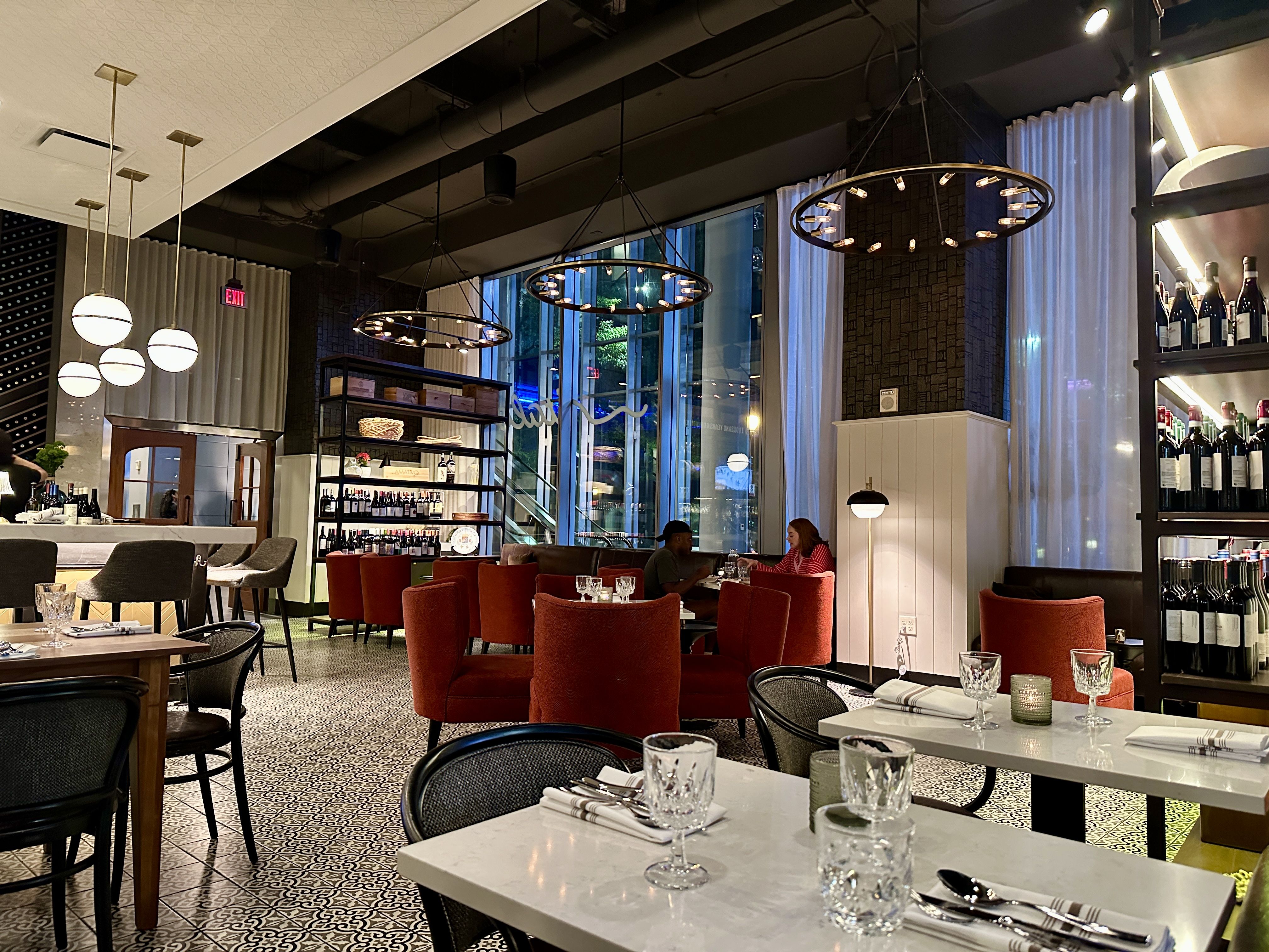 italo uptown charlotte. Modern restaurant interior with white marble tables, red upholstery, and glassware. Large circular pendant lights, exposed ceiling beams, patterned tile floor, and wine shelves by tall windows.