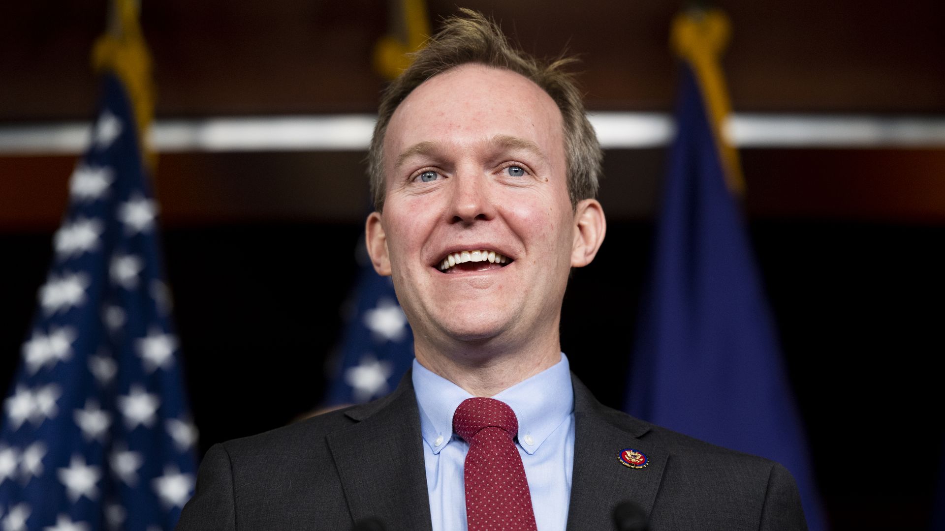 Smiling man in dark suit, light blue shirt, and red tie speaking at a podium with two American flags in the background.