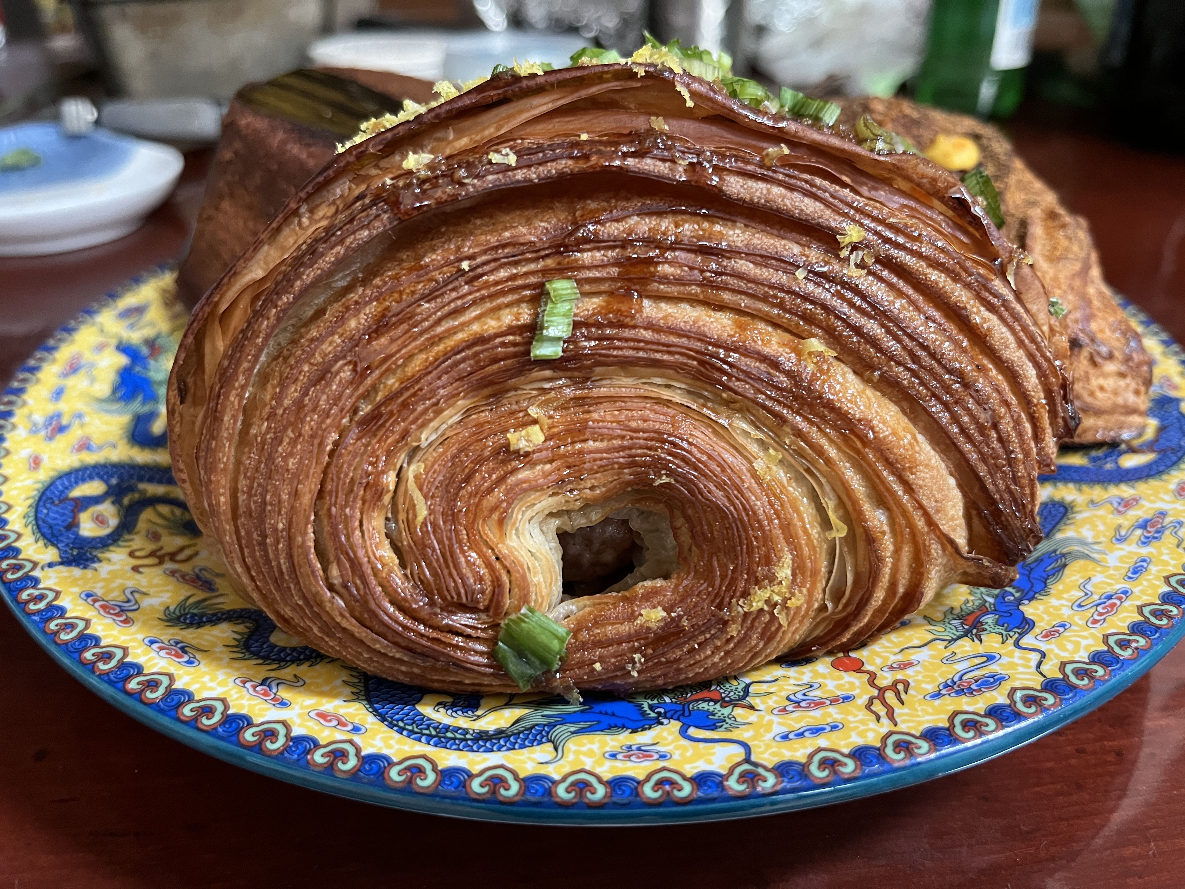 Close-up of a crispy, layered golden-brown onion or pastry topped with chopped green onions, served on a colorful blue and yellow plate with dragon designs on a brown table.