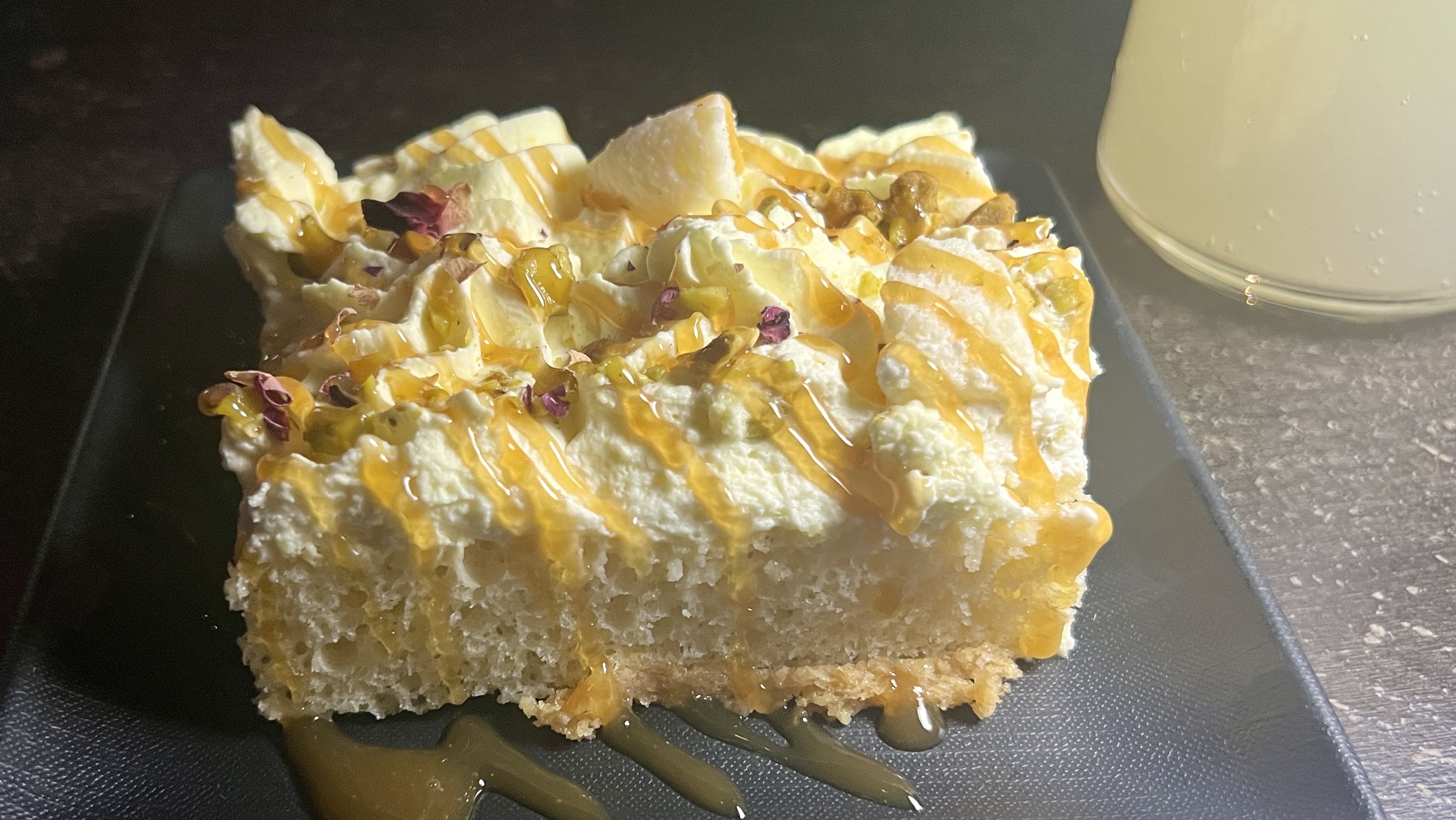 A rectangular slice of cake with yellow drizzling and pistachio crumble