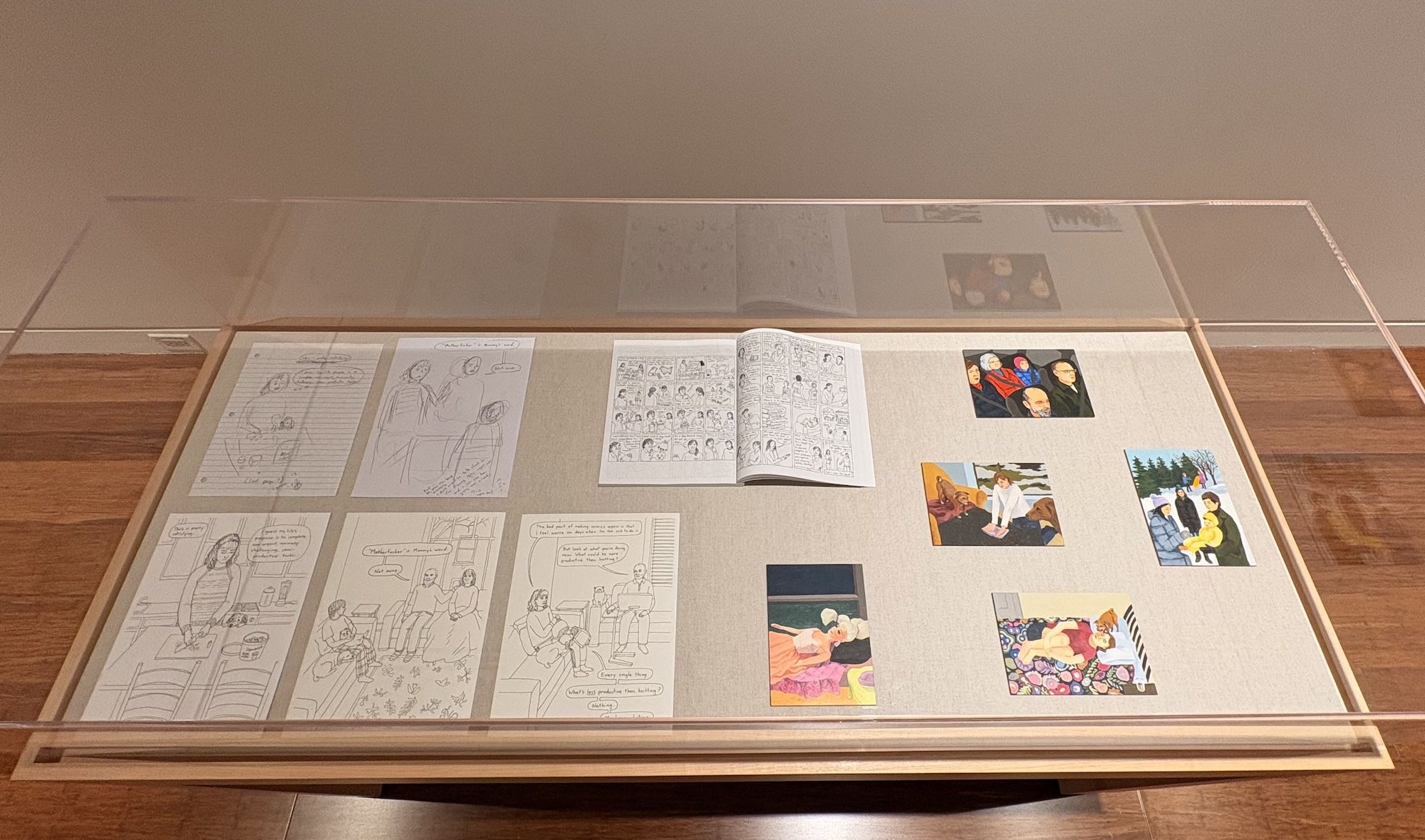 Display case with a glass top contains multiple comic pages and color panels laid out on a light gray mat; an open comic book sits center, showing drawn scenes of people.