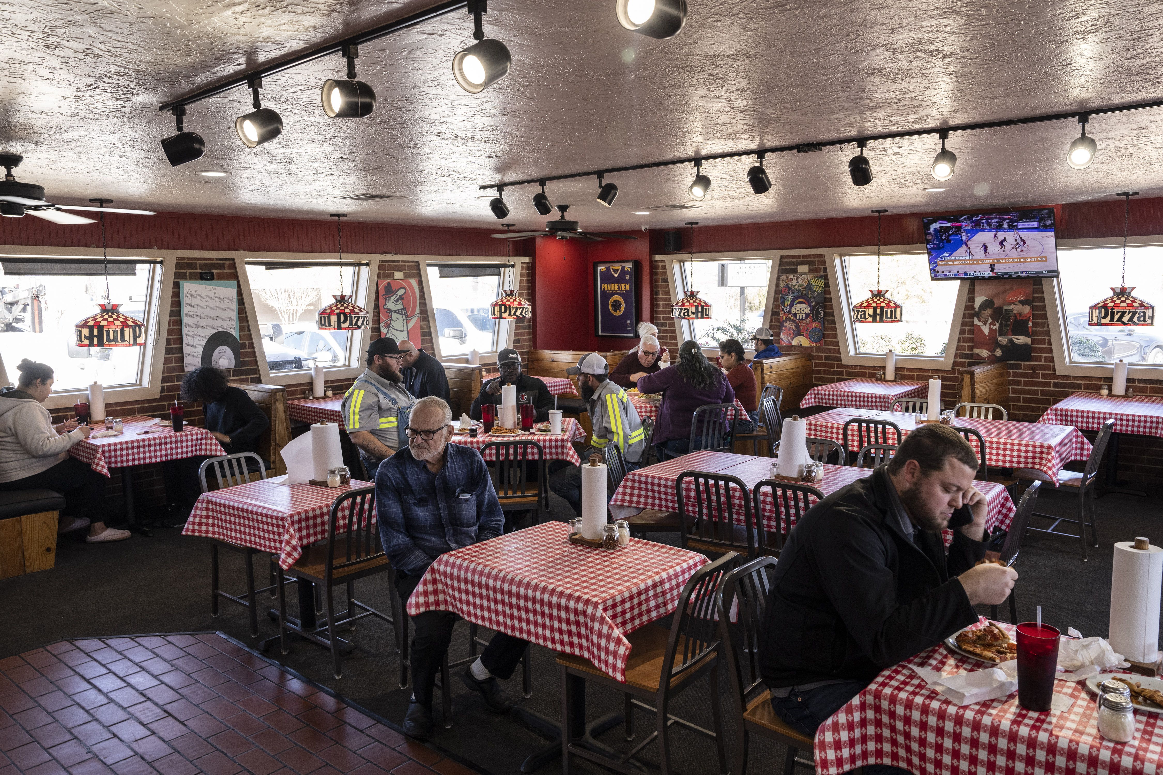 A retro Pizza Hut interior with red brick walls and red-and-white checkered tables. Diners sit at booths and tables; a man on the right eats pizza while on his phone; a TV shows sports.