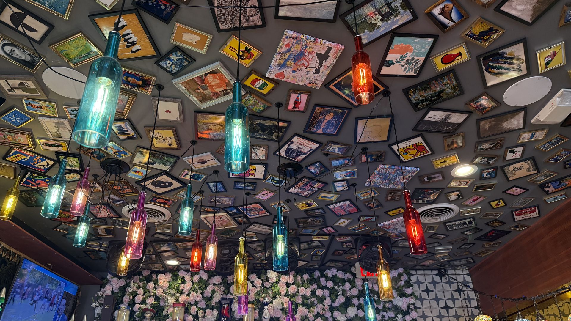 Ceiling covered with numerous framed pictures and paintings, colorful bottle-shaped lights hanging down, and a wall decorated with pink and white flowers in a cozy indoor setting.