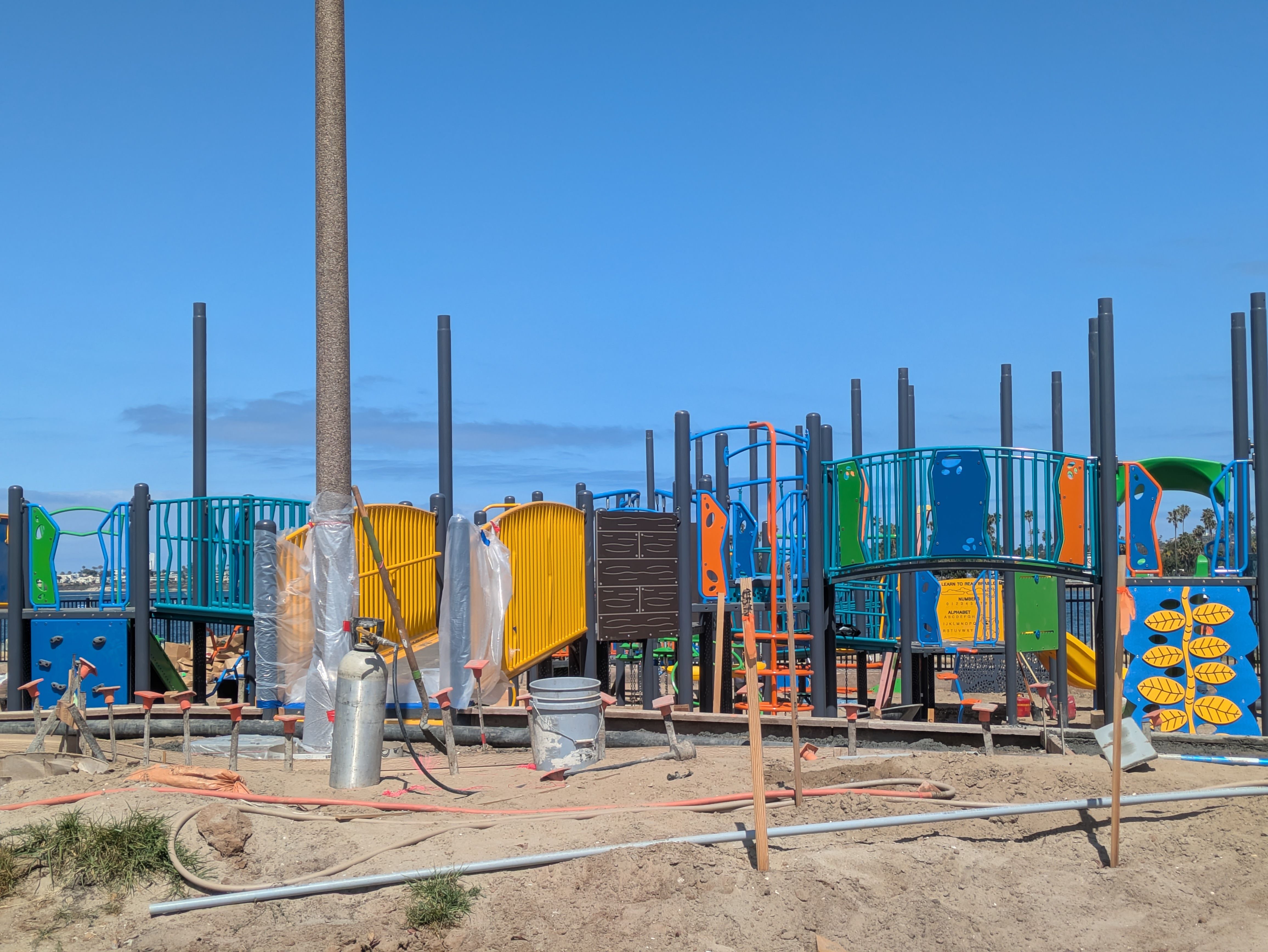 Under-construction playground with bright blue, orange, and green panels, many vertical gray posts, and exposed dirt under a clear blue sky.