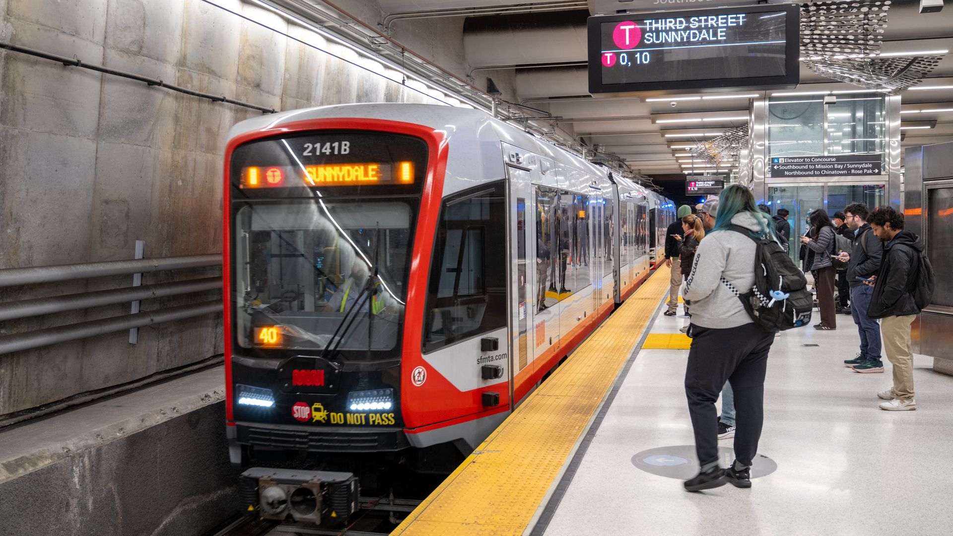 Underground metro platform with a red-and-white train at the edge; its front display reads "Sunnydale". A digital sign above shows "Southbound" and "Third Street Sunnydale". Passengers wait to the right.