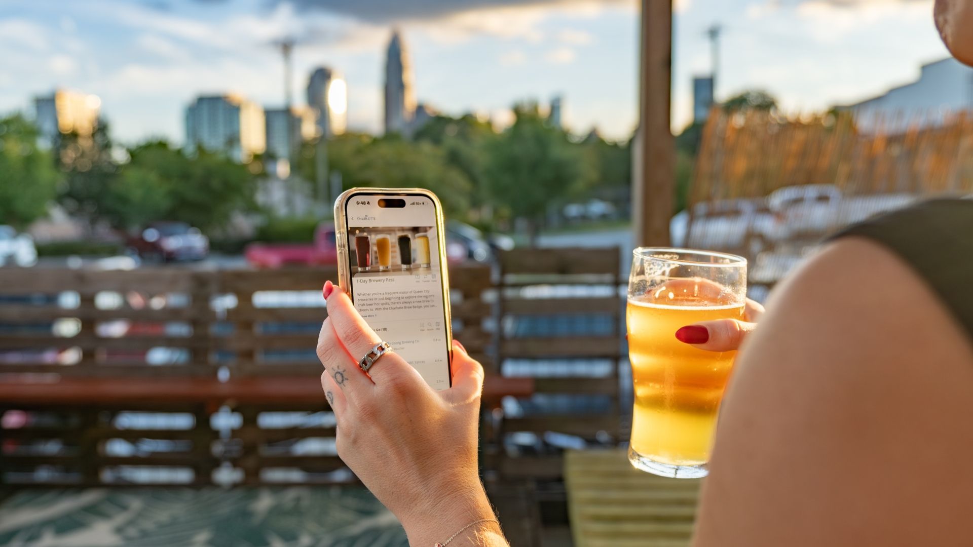 Person with red nail polish holding a glass of beer in one hand and a smartphone displaying a 1-Day Brewery Pass app screen with images of beer on a sunny patio with a city skyline background.