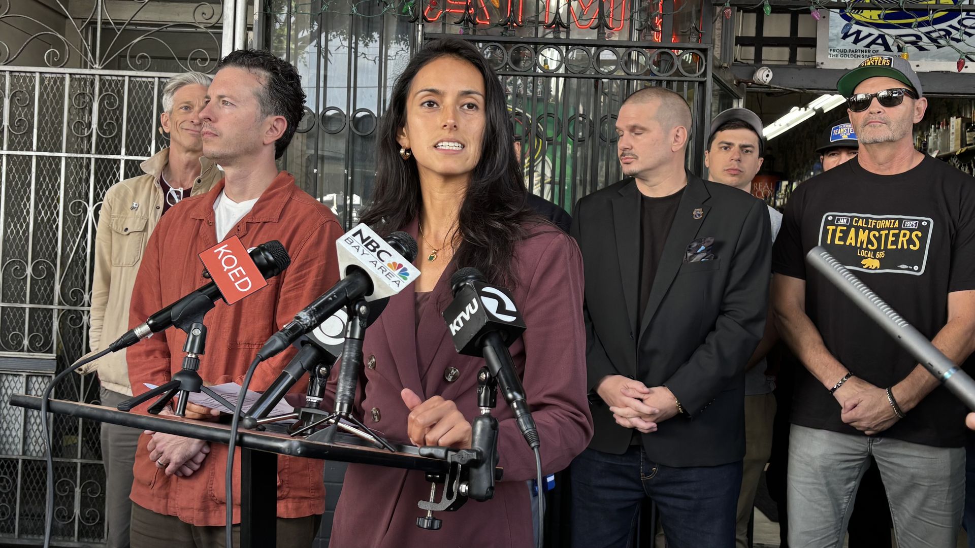 Woman in a maroon blazer speaks at a street press conference with microphones labeled NBC Bay Area, KOED, and KTVU, as several men stand behind her near a gated storefront.
