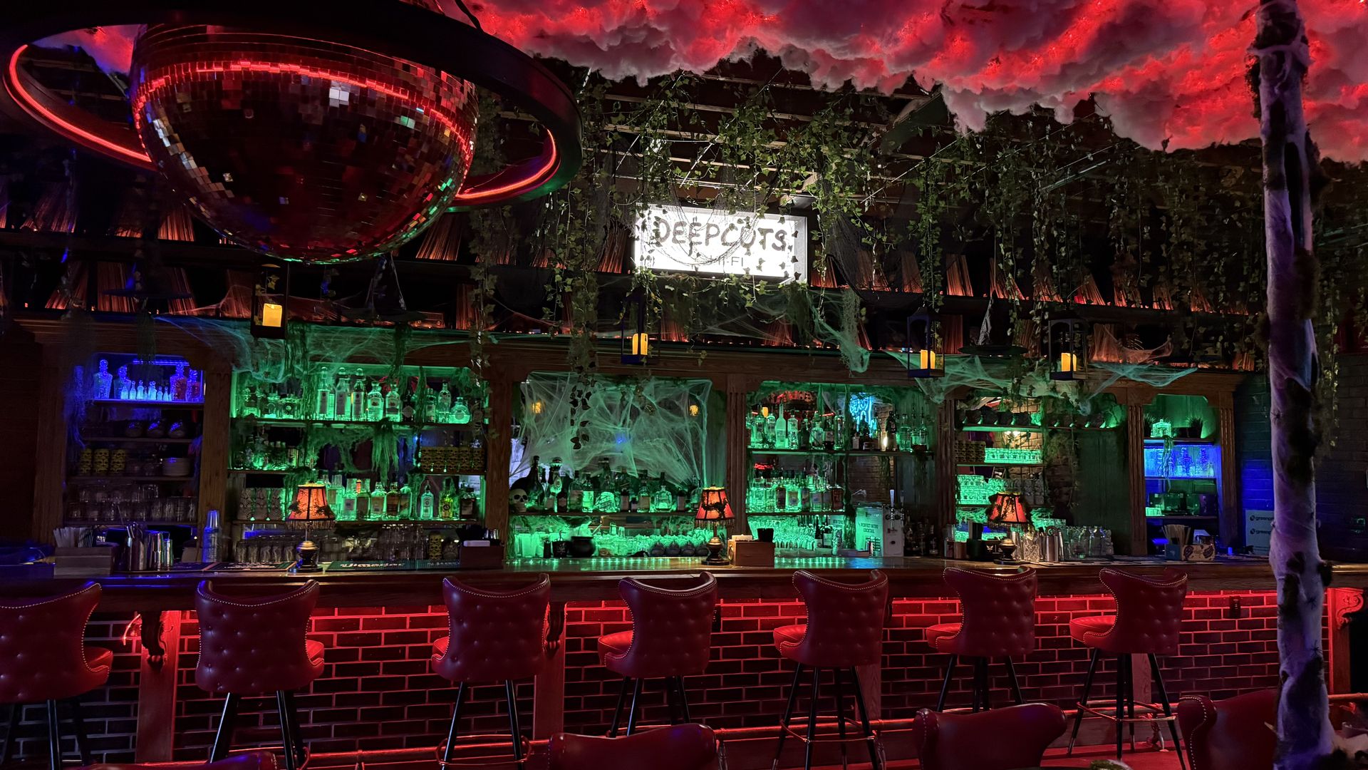 Neon-lit bar interior with a large hanging disco ball casting red reflections; green-lit bottles behind the bar, plus fake cobwebs and hanging vines across the ceiling.