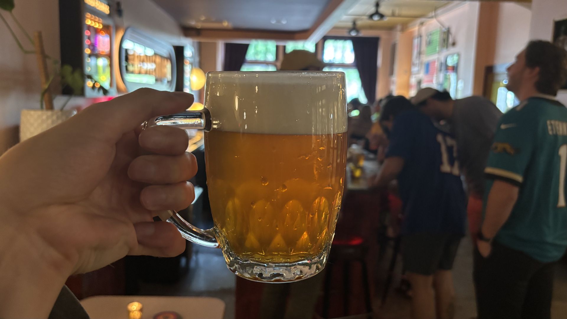 Hand holding a large glass beer mug with a foamy white head in a crowded bar; blurry patrons at a counter, neon signs and screens in the background.