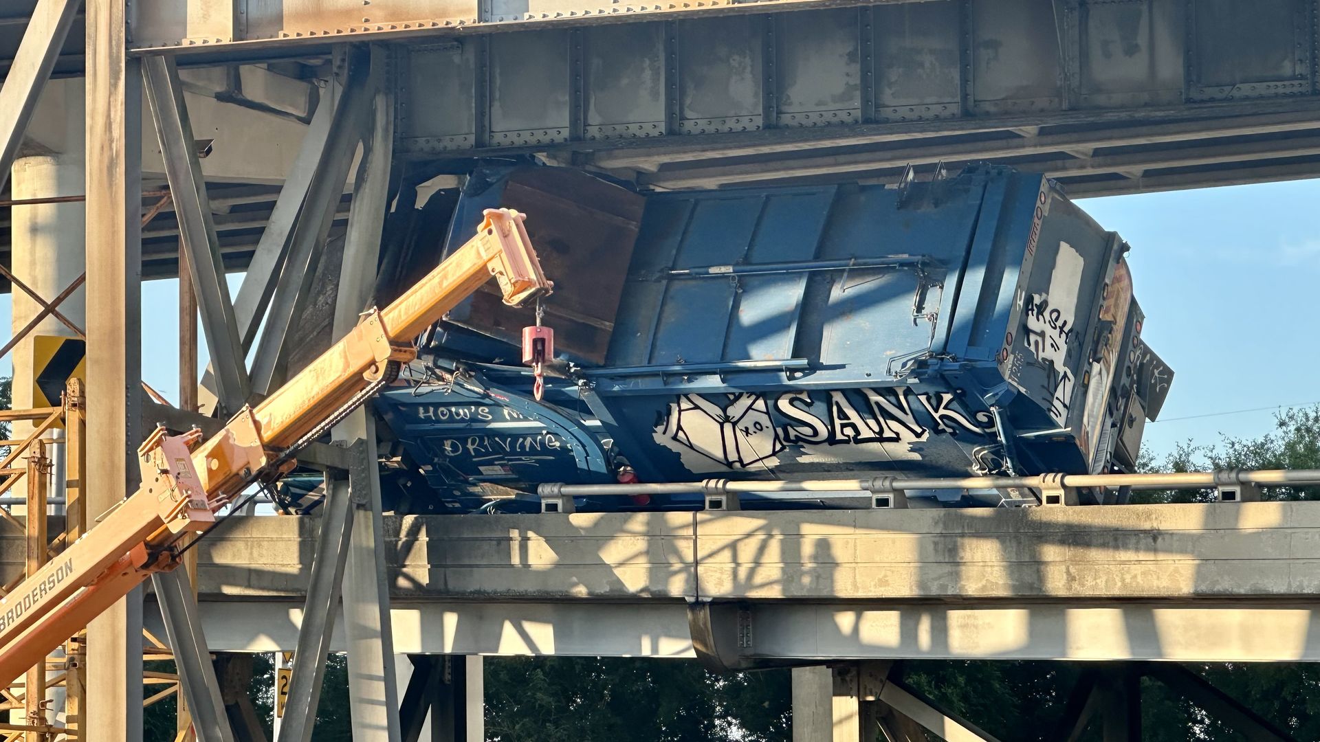 A blue garbage truck is wedged under an interstate overpass, its rear end lifted and damaged. A crane is positioned nearby, assisting with the removal. The truck has graffiti and visible damage, and the scene is framed by steel support beams and concrete infrastructure.