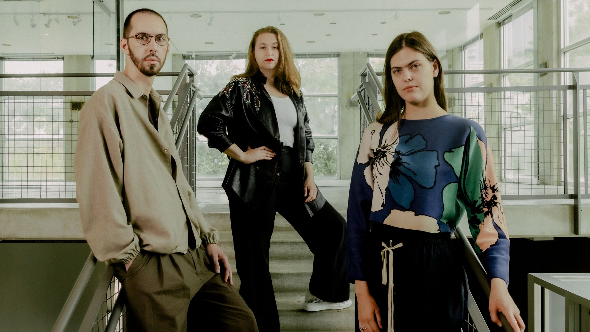 Three young adults standing on a modern stairway with metal railings, large windows in background. Man on left wears glasses, beige shirt, and brown pants. Middle woman in black jacket and pants. Woman on right in colorful floral top and black skirt.