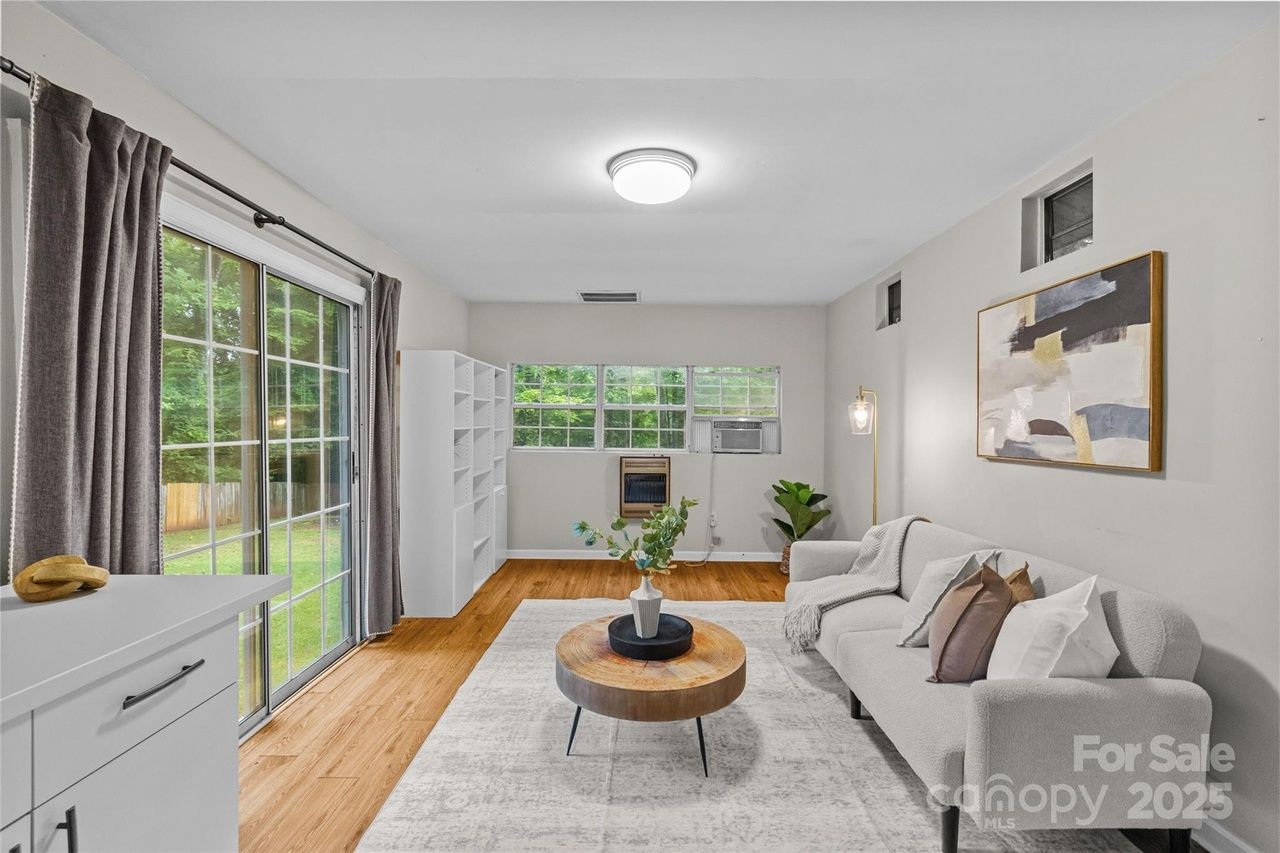 Bright living room with light gray sofa, abstract wall art, large windows with gray curtains, wood floor, round wooden coffee table, white shelving unit, and sliding glass door to backyard.