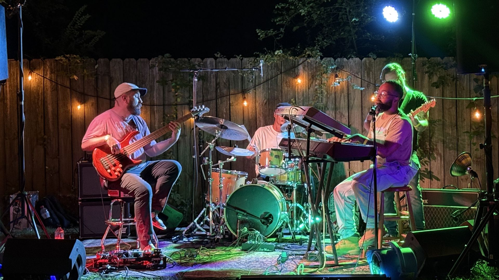 Four musicians playing bass, drums, keyboard, and guitar outdoors at night with colorful stage lights and string lights against a wooden fence backdrop.