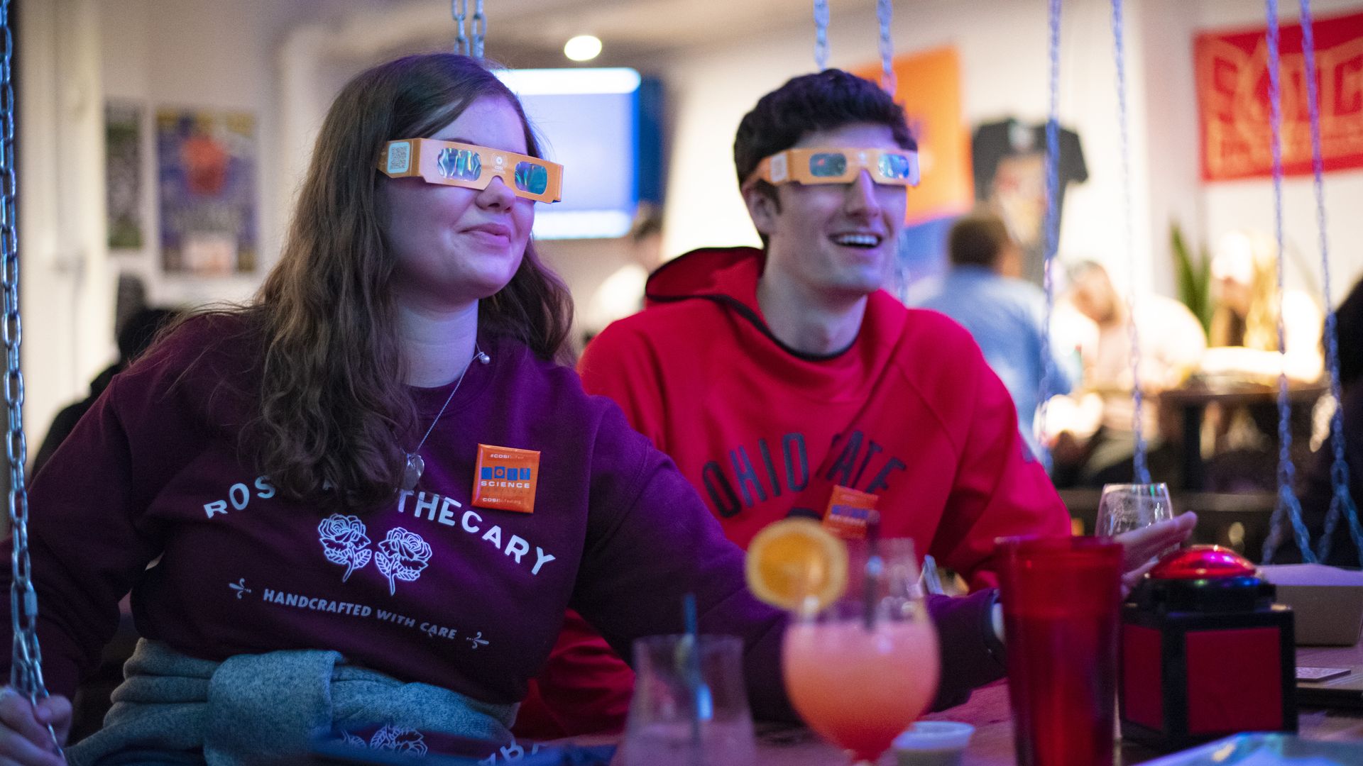 Two people wearing 3D glasses at a bar press a trivia buzzer