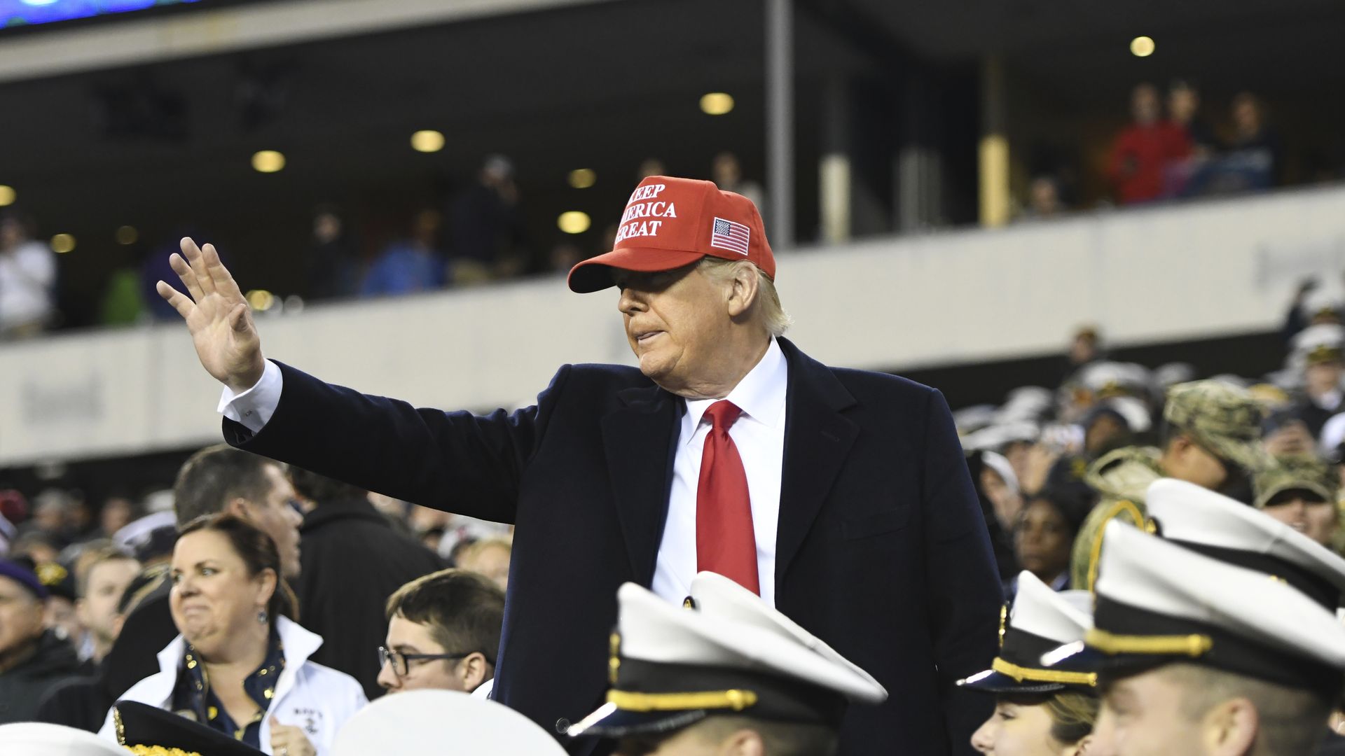 In this image, Trump stands in a MAGA hat amid people wearing Navy hats and uniforms