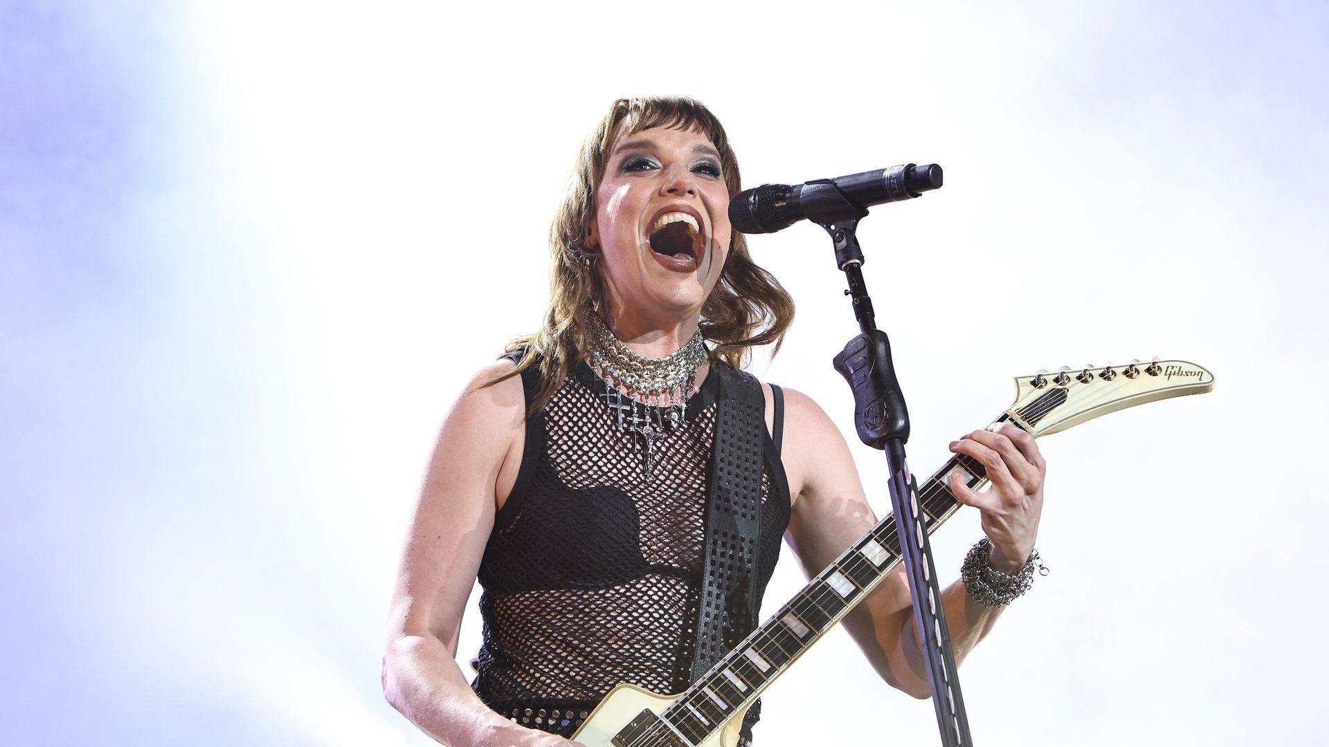 A female rock singer with short hair and smoky makeup belts into a microphone while playing a cream Gibson electric guitar on a bright stage, wearing a black mesh top and layered jewelry.