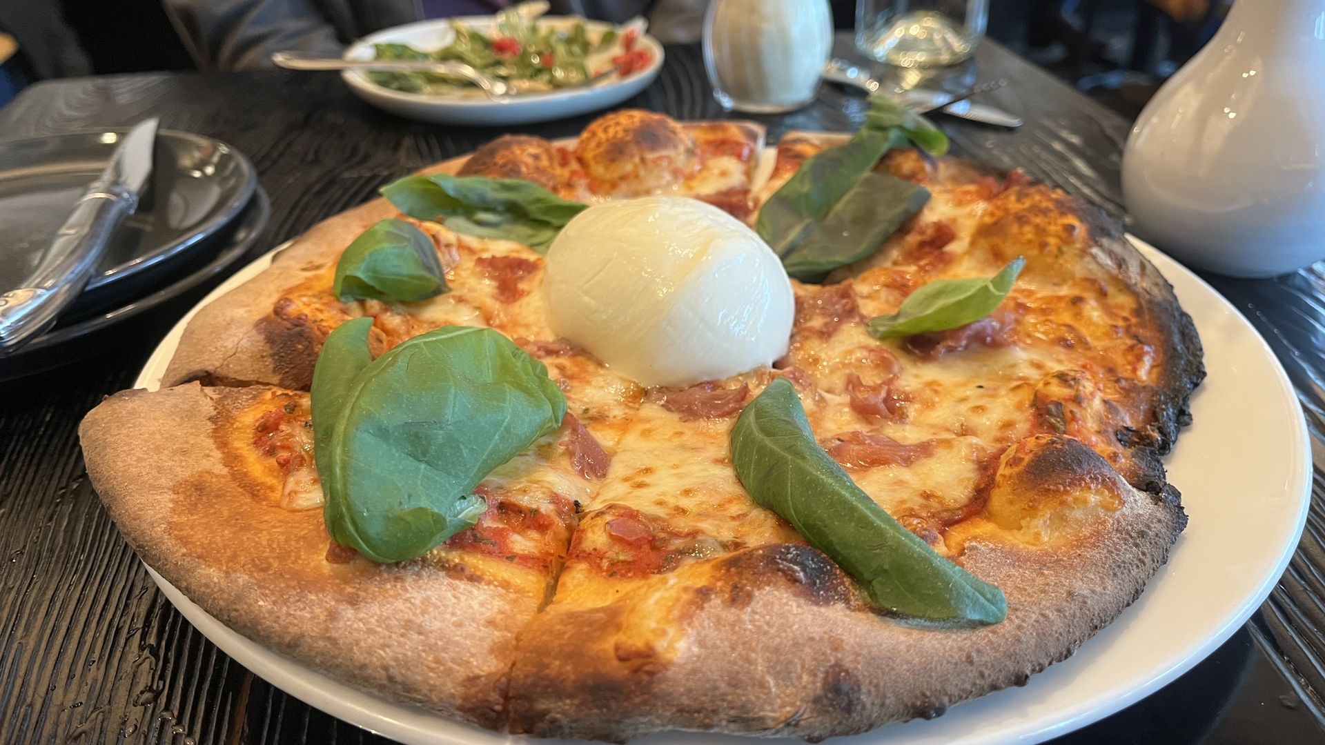 Pizza on a white plate with a thick, crispy crust, melted cheese, and fresh basil leaves. A large ball of mozzarella sits in the center; a wooden table, salad plate, and pitcher in the background.