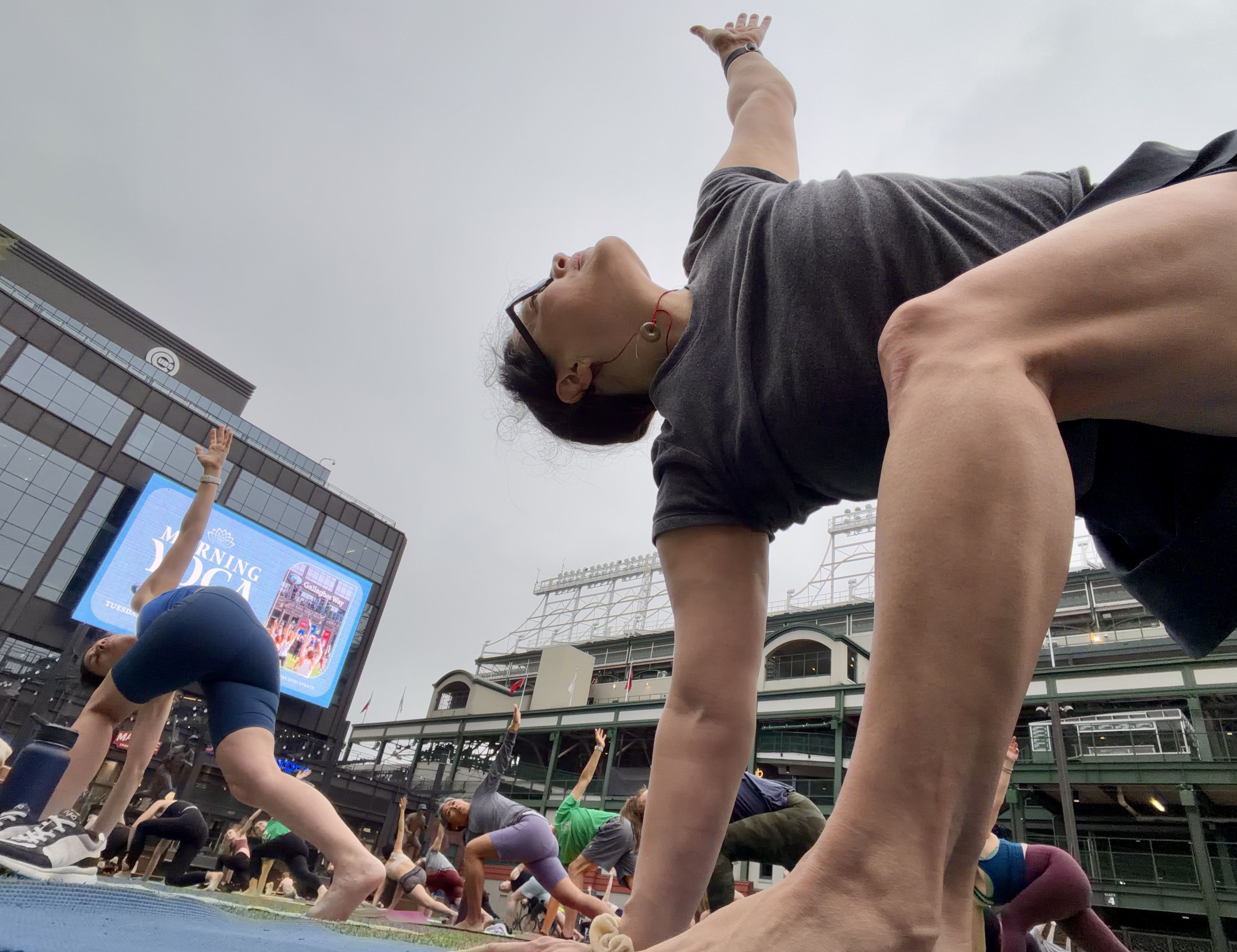 Outdoor group yoga class in a city plaza; dozens of people in athletic wear twist and reach with arms raised. A tall glass building and a large digital billboard loom in the background under a gray sky.