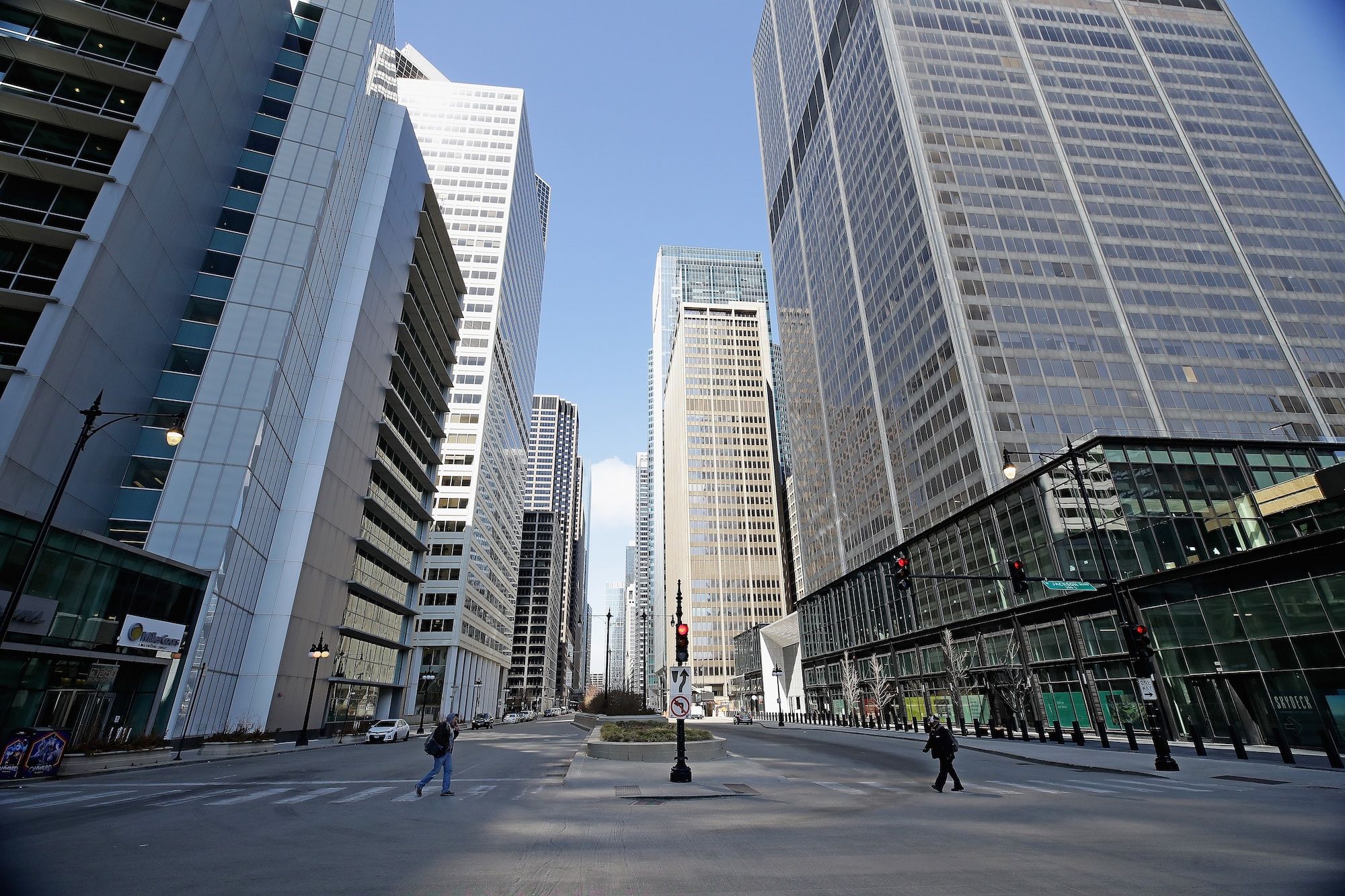 Office buildings line a nearly empty city street with two people crossing the street.