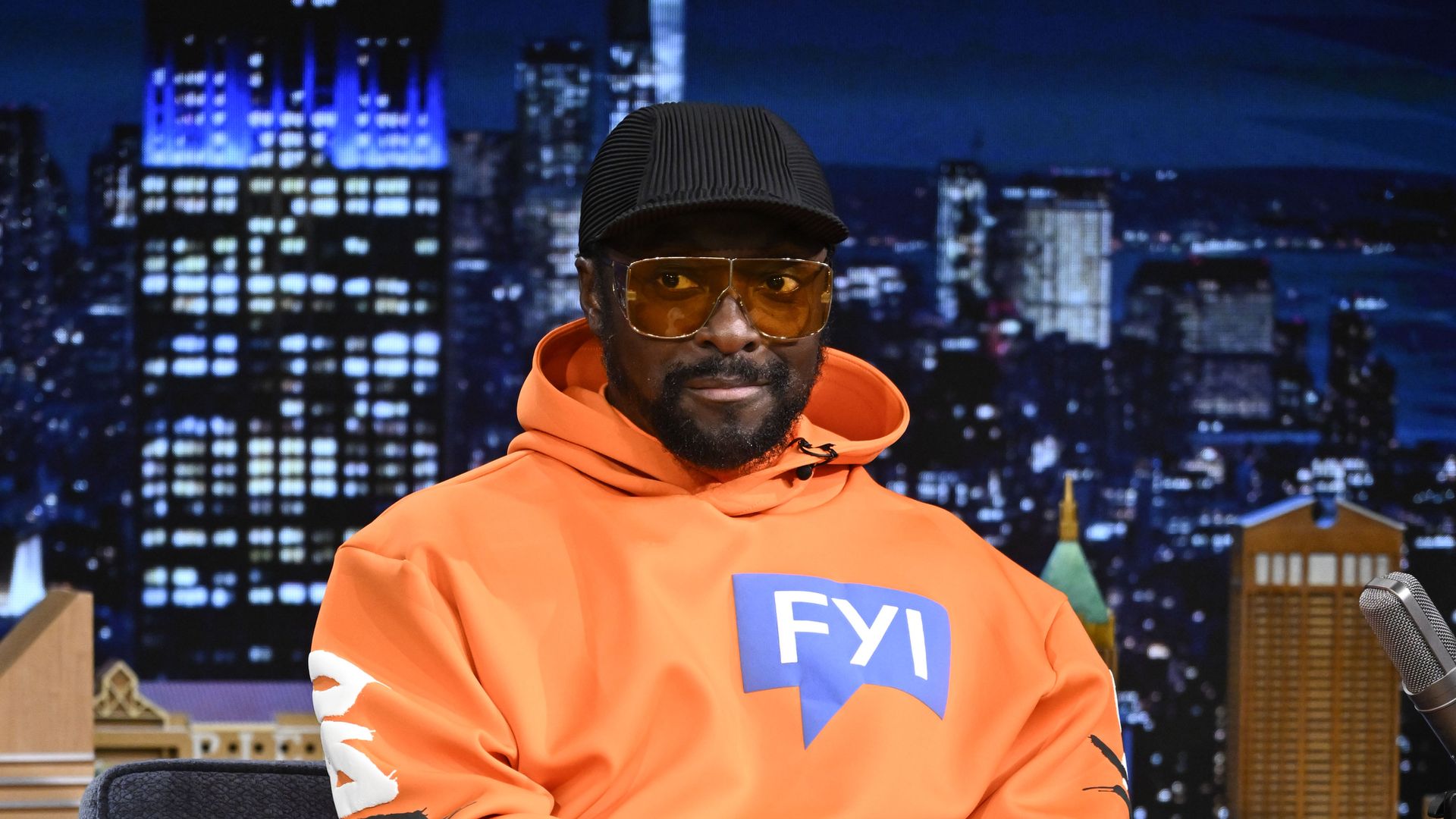 Will.i.am on The Tonight Show Starring Jimmy Fallon wearing a bright orange sweatshirt that says FYI, a black hat and sunglasses