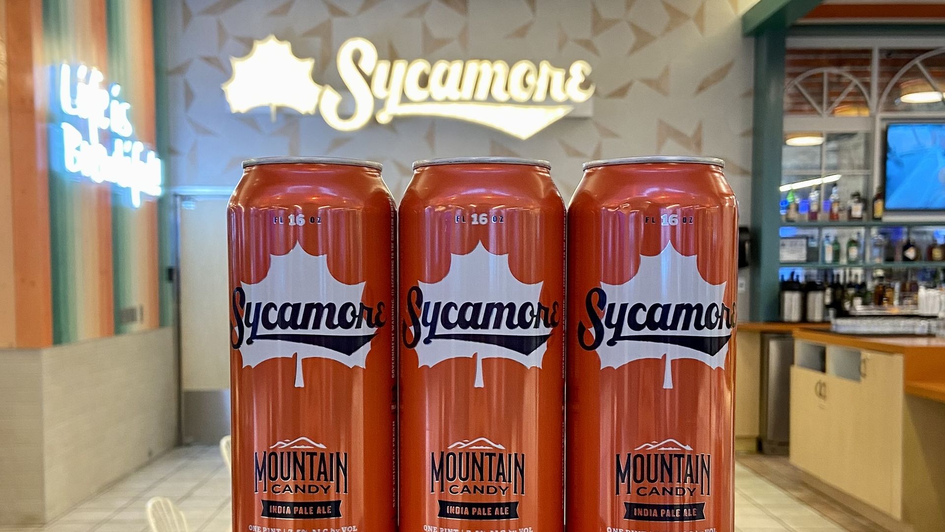 Seven orange cans of Sycamore Mountain Candy India Pale Ale stacked in a pyramid on a bar counter in a bright, modern bar with a lit Sycamore sign in the background.