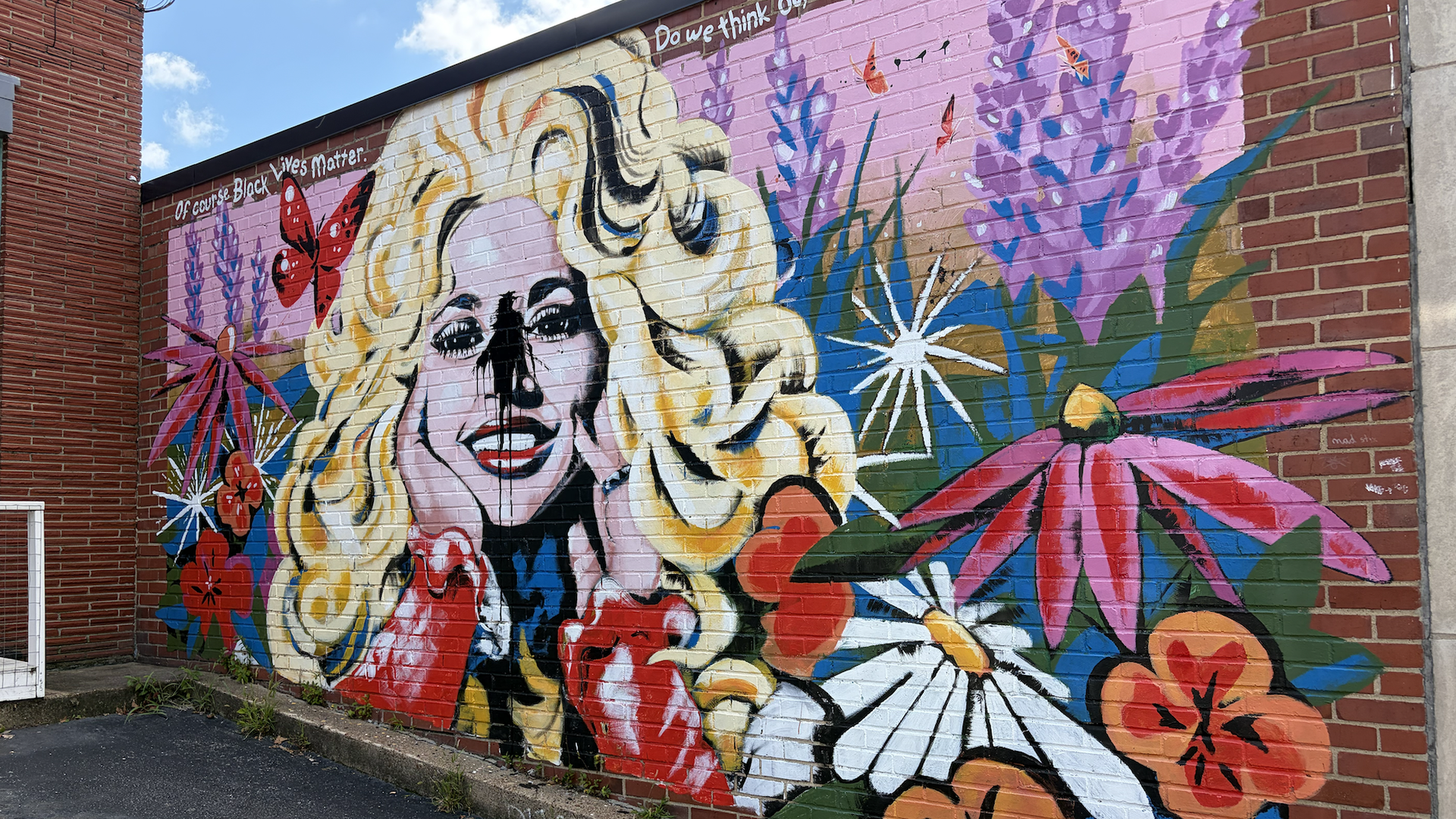 Bright mural on a brick wall depicting a stylized blonde woman with flowing hair, surrounded by flowers and butterflies on a pink and purple background; text reads "Of course Black Lives matter."