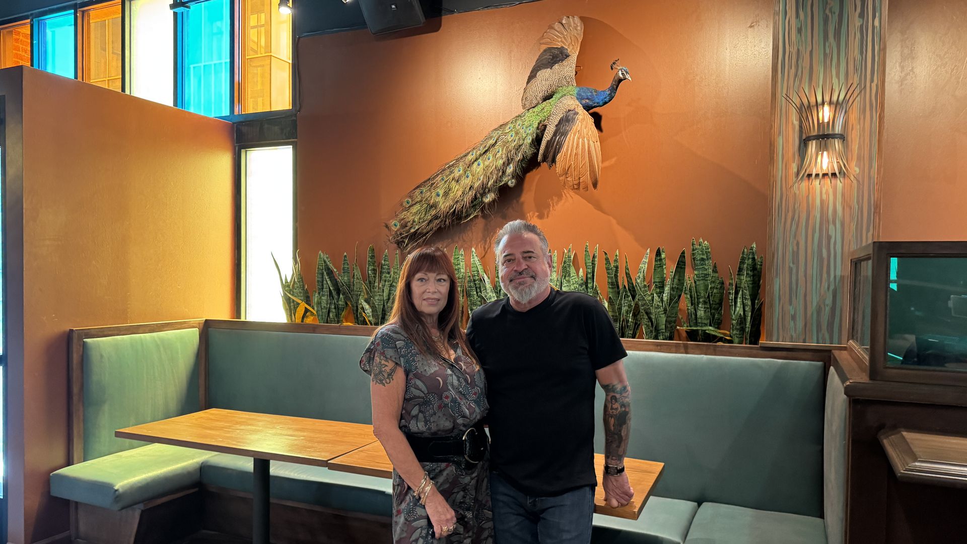 A man and woman stand in a restaurant booth with green seating, orange walls, plants, and a colorful stained glass window. A large mounted peacock spreads its wings above them.