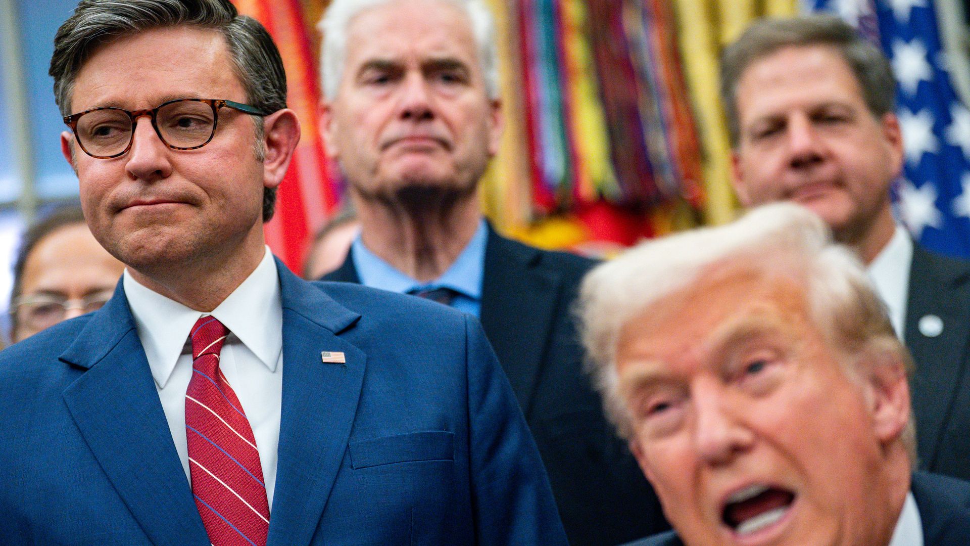 A man in a blue suit with glasses and a red striped tie looks down with a neutral expression while a man with blond hair in the foreground speaks, with colorful flags and blurred men in suits behind them.