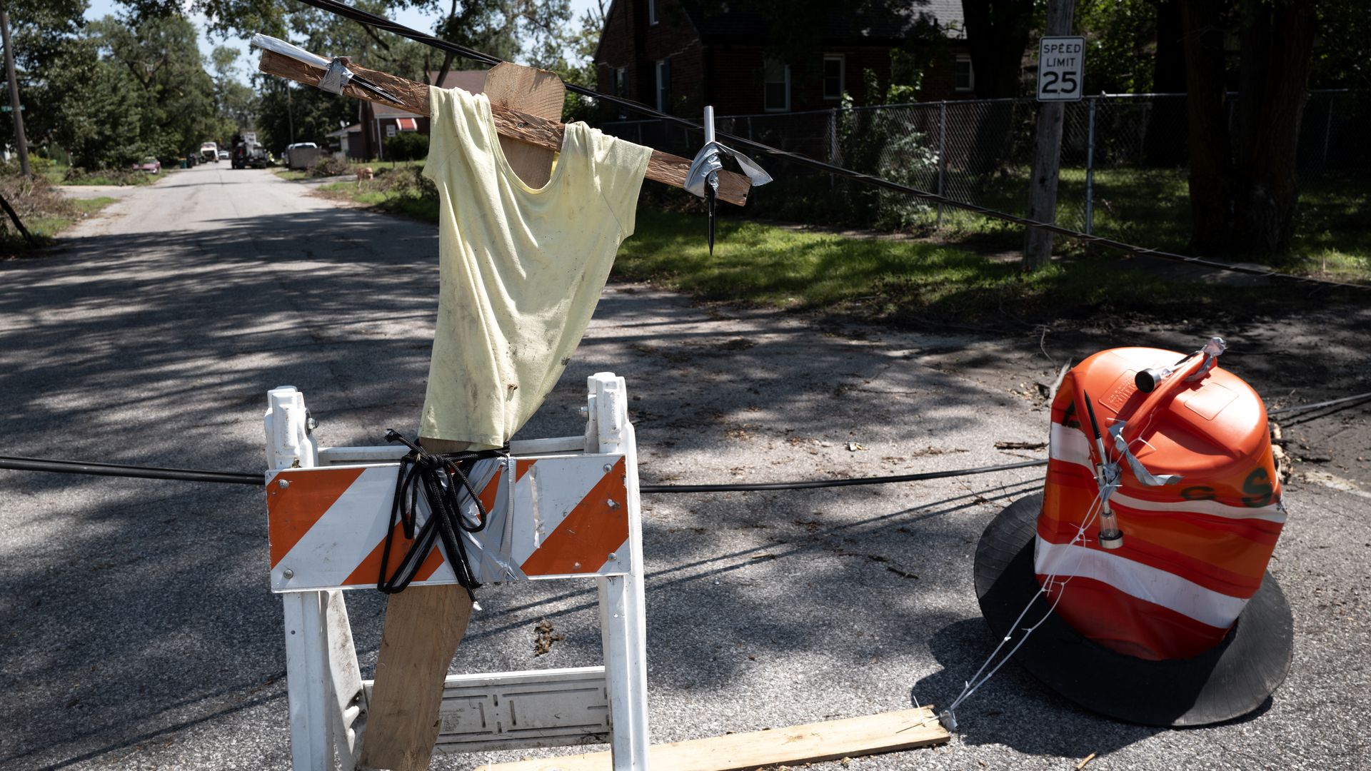 Residential street blocked by orange barricades. A pale yellow shirt drapes over a wooden crossbeam tied to power lines, forming a crude figure. A 25 mph sign and fence in the background.