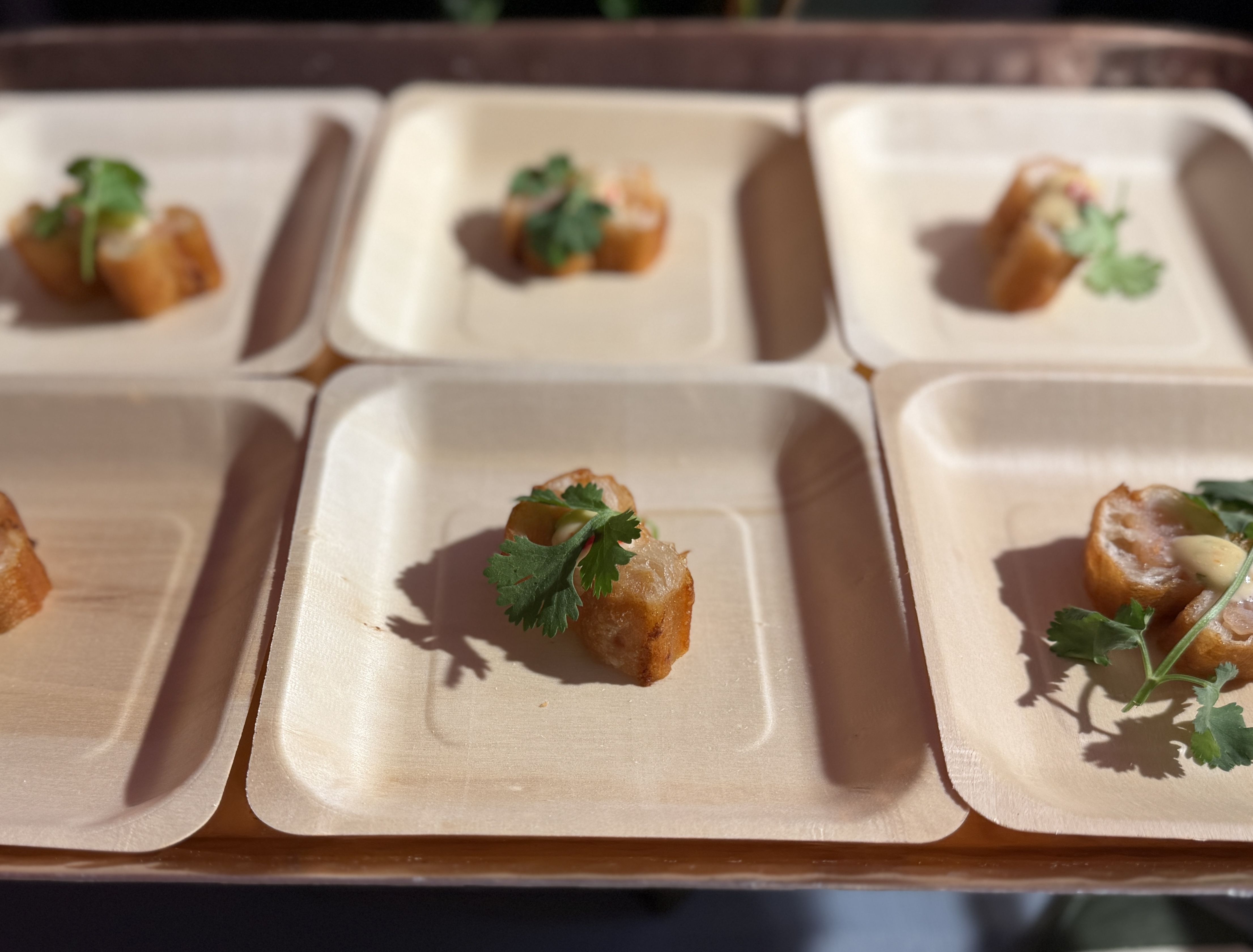 Blurry image of several small fried bite-size appetizers in a white foam tray, each topped with a cilantro leaf and greens, arranged in individual compartments.