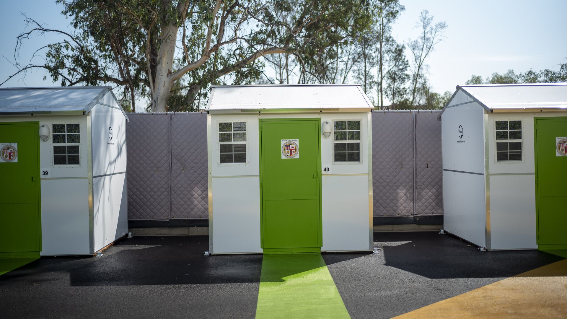 A row of three white tiny homes with green doors in front of a fence with a tree in the background.