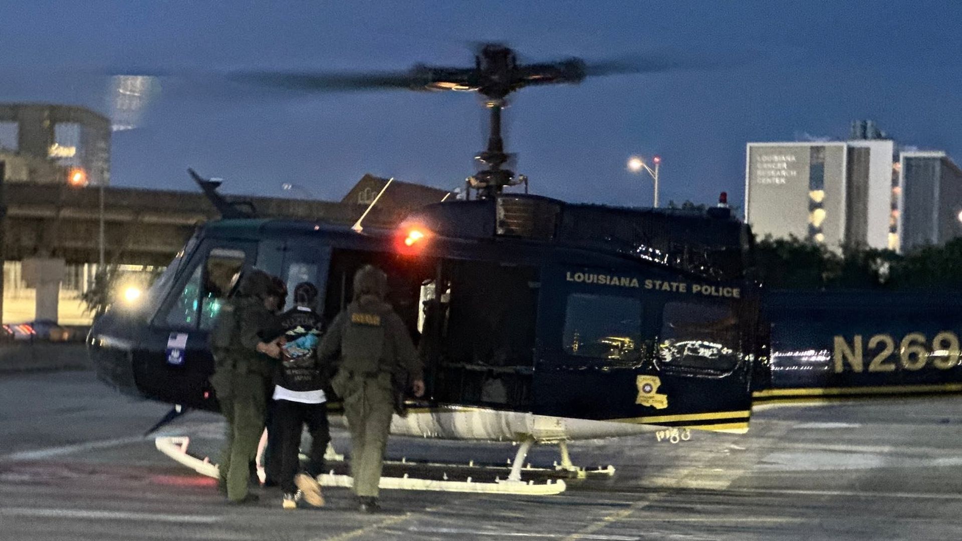 Three individuals, dressed in dark uniforms, escort a fourth person towards a black helicopter with 'Louisiana State Police' printed on its side. The scene appears to be on a tarmac, and the helicopter’s rotor blades are blurred, indicating motion. The photograph is taken in low light, possibly duri