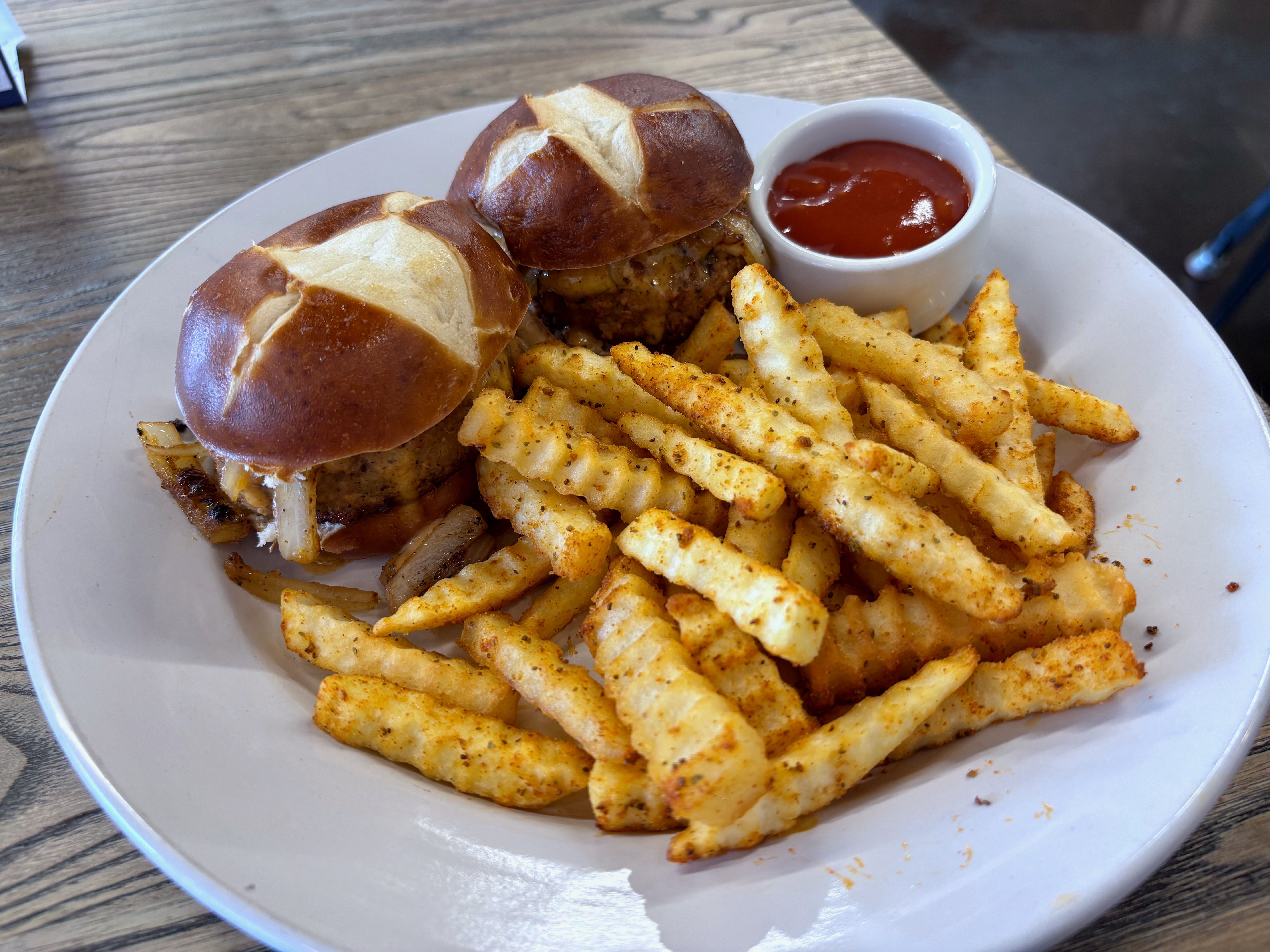 Plate with two burgers on pretzel buns, melted cheese and onions visible, beside a pile of seasoned crinkle-cut fries and a small cup of ketchup on a wooden table.
