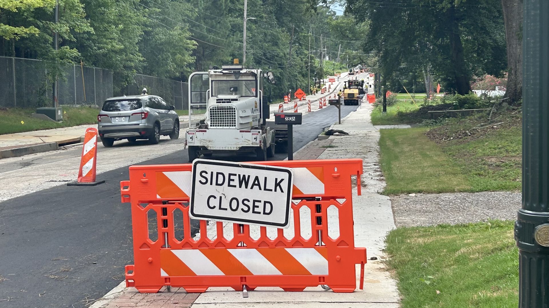 Orange barrier with "SIDEWALK CLOSED" sign blocking sidewalk near road construction with machinery and orange cones on a tree-lined residential street.
