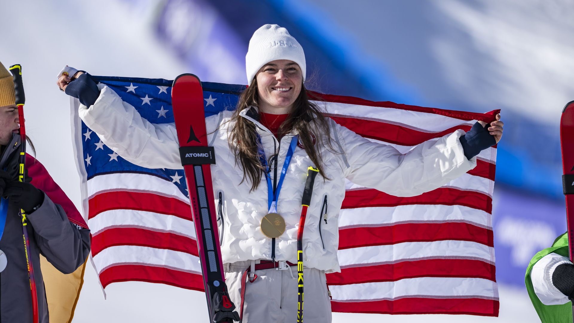 Smiling skier in white jacket and beanie holding an American flag, wearing a gold medal, with red and black skis, standing on snow at a ski competition podium.