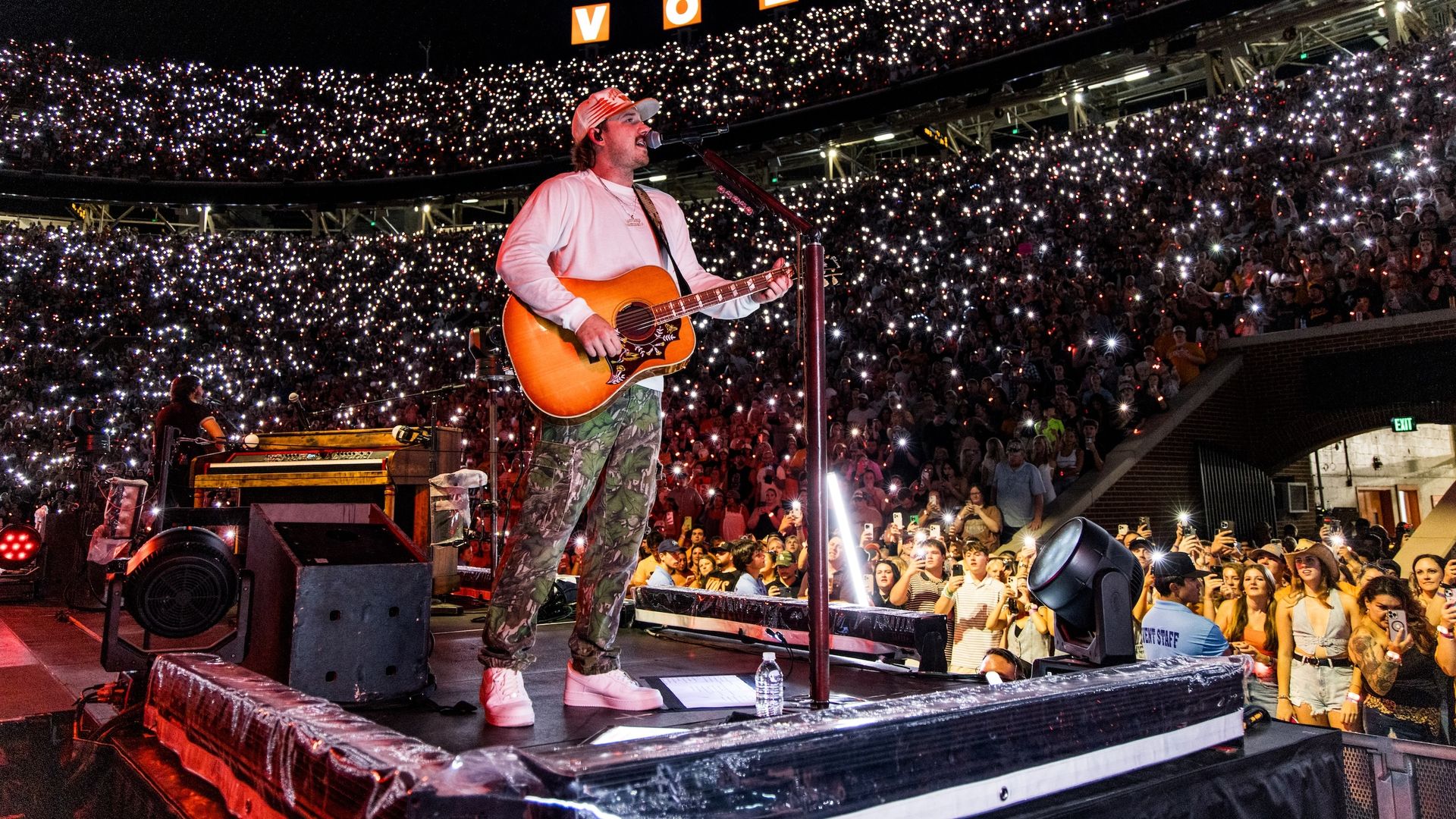 Morgan Wallen in camouflage pants and white shirt plays guitar on stage at night, with thousands of audience members holding up lights in a stadium with "VOLS" sign illuminated.
