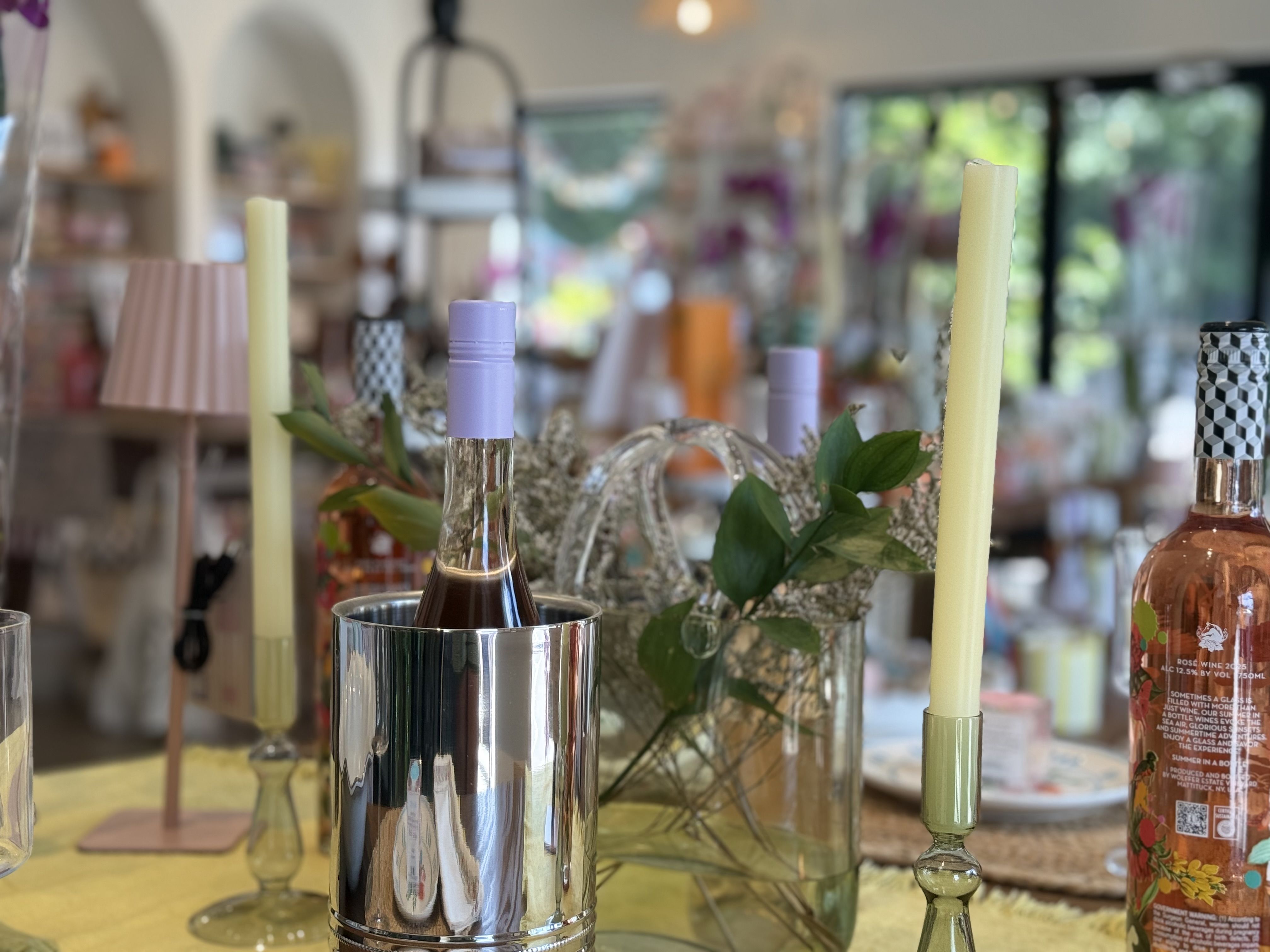 Close-up of a shop display: tall pastel candles in shiny metal cups, lavender bottle with purple cap, glass vases, greenery, and a blurred colorful background.
