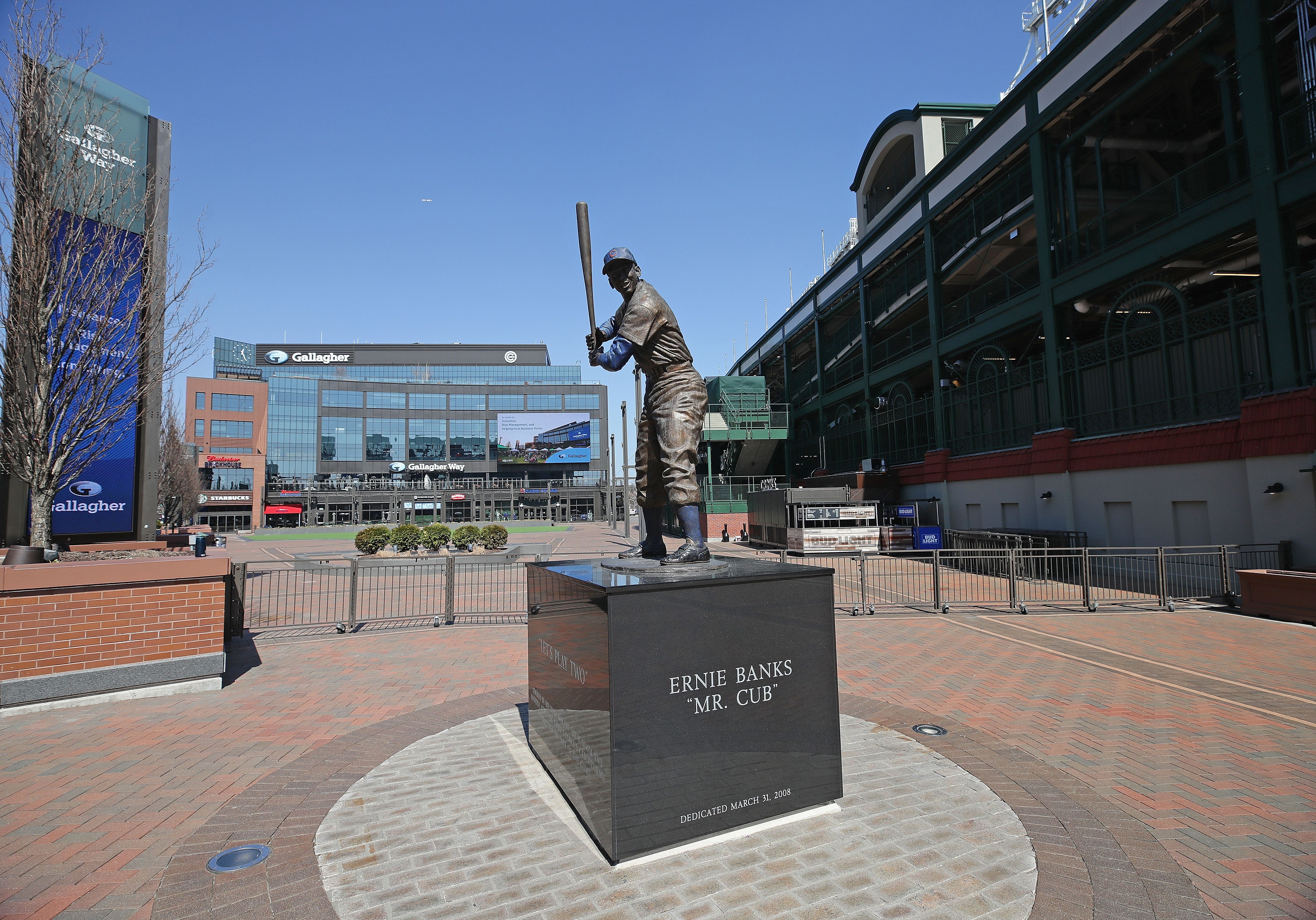 Statue of baseball player Ernie Banks holding a bat, standing on a black pedestal reading "Ernie Banks 'Mr. Cub' dedicated March 31, 2008" outside a stadium under a clear blue sky.