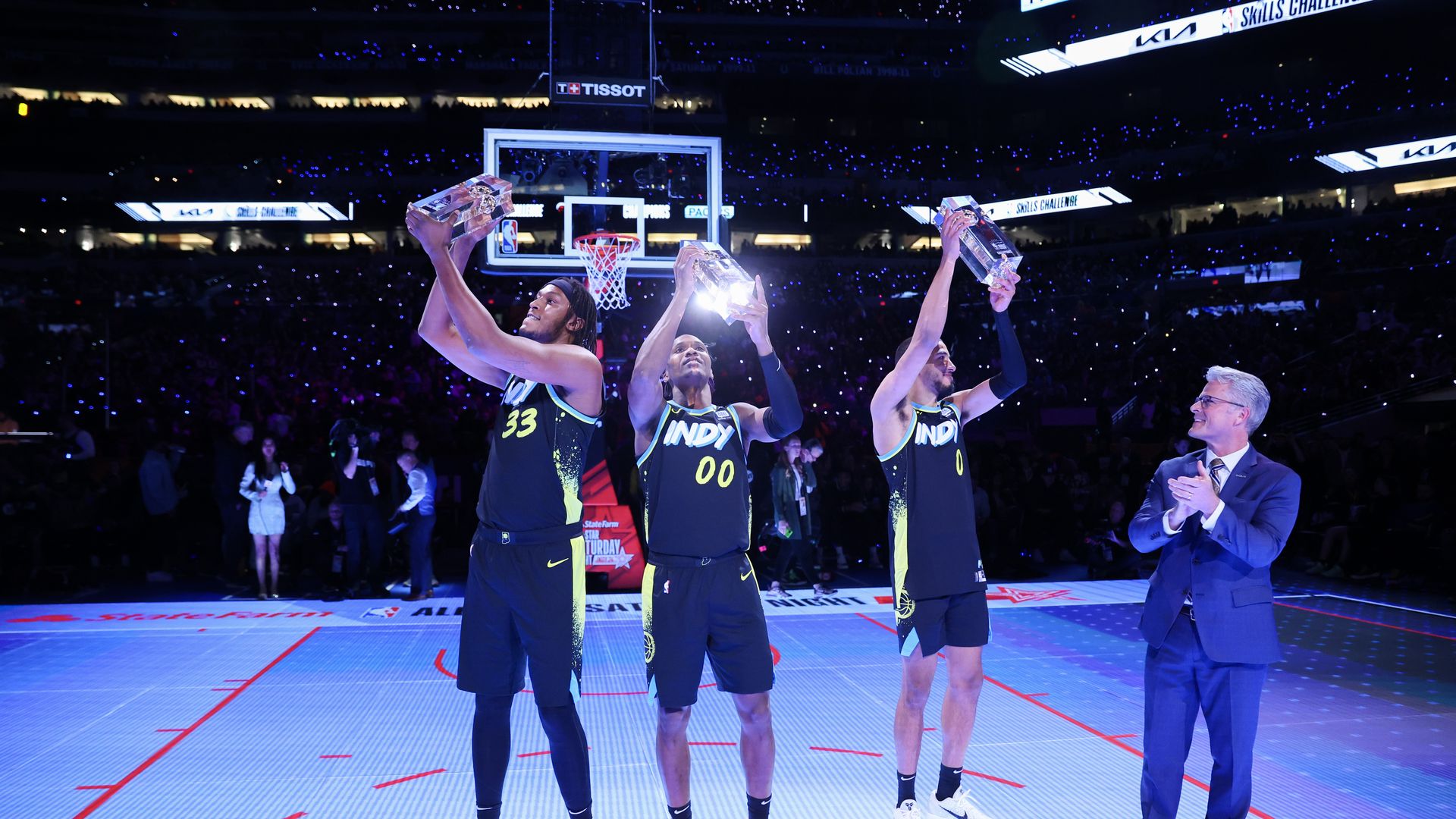 Three basketball players on a court holding trophies.